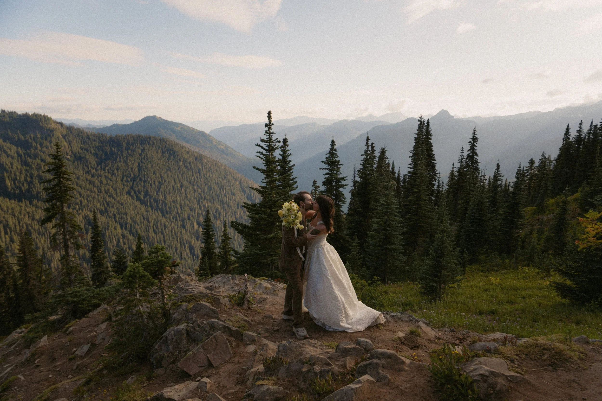 A bride and groom sharing a kiss on a rocky mountaintop surrounded by pine trees with mountains in the background during sunset or sunrise.
