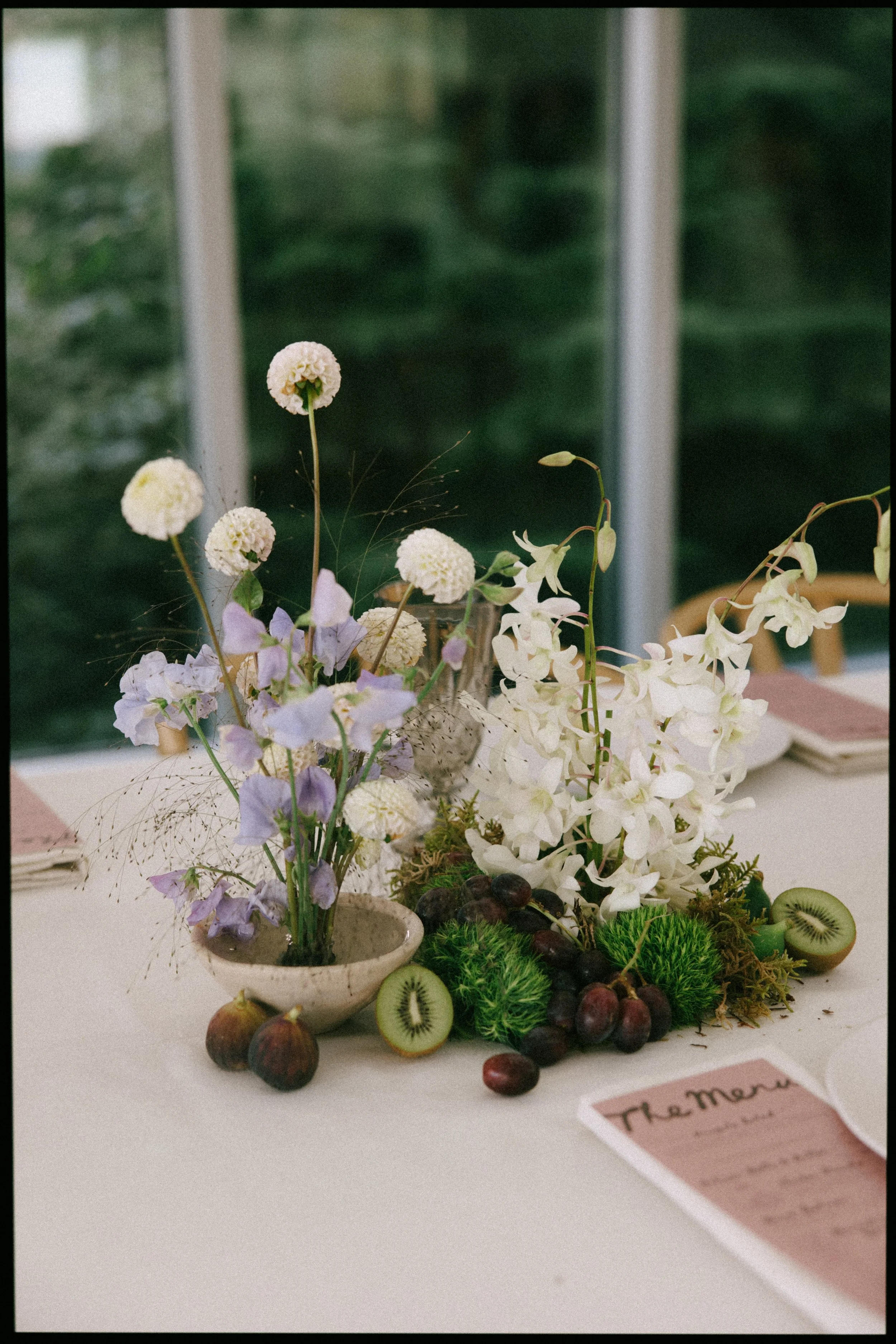 A table centerpiece with white and light purple flowers, green moss, kiwi slices, and dark grapes, with a partially visible menu on the right side.