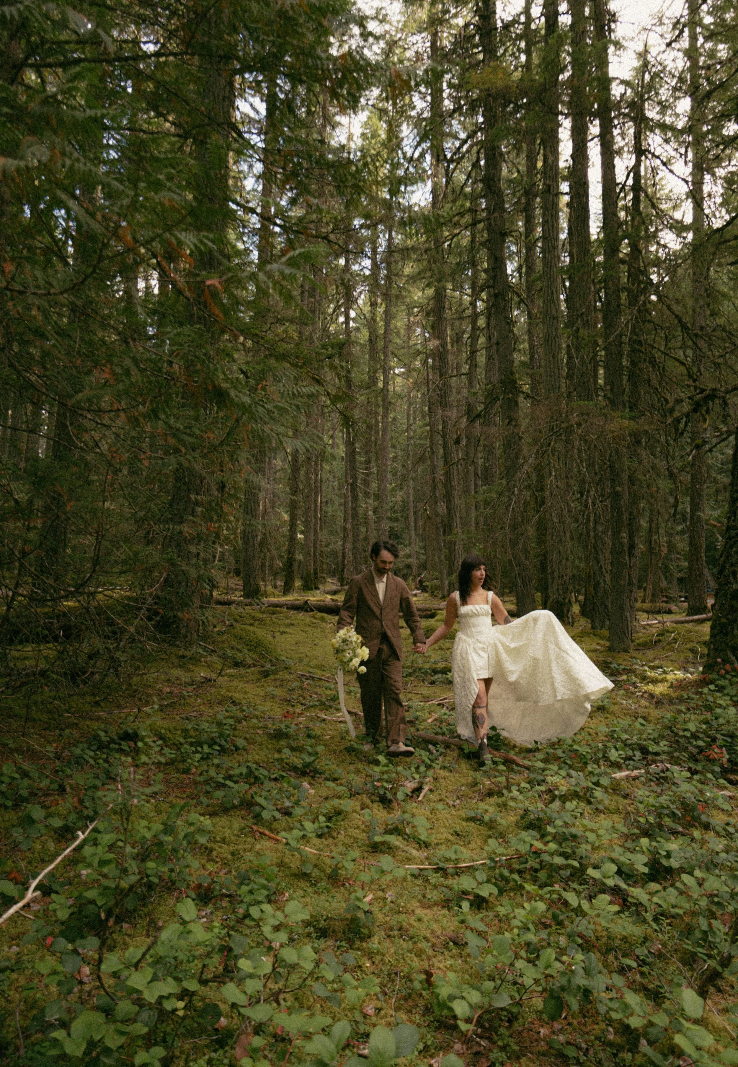 A couple walking hand-in-hand in a forest trail, with the woman lifting her cream-colored dress to walk over mossy ground, and the man carrying a bouquet of white flowers.