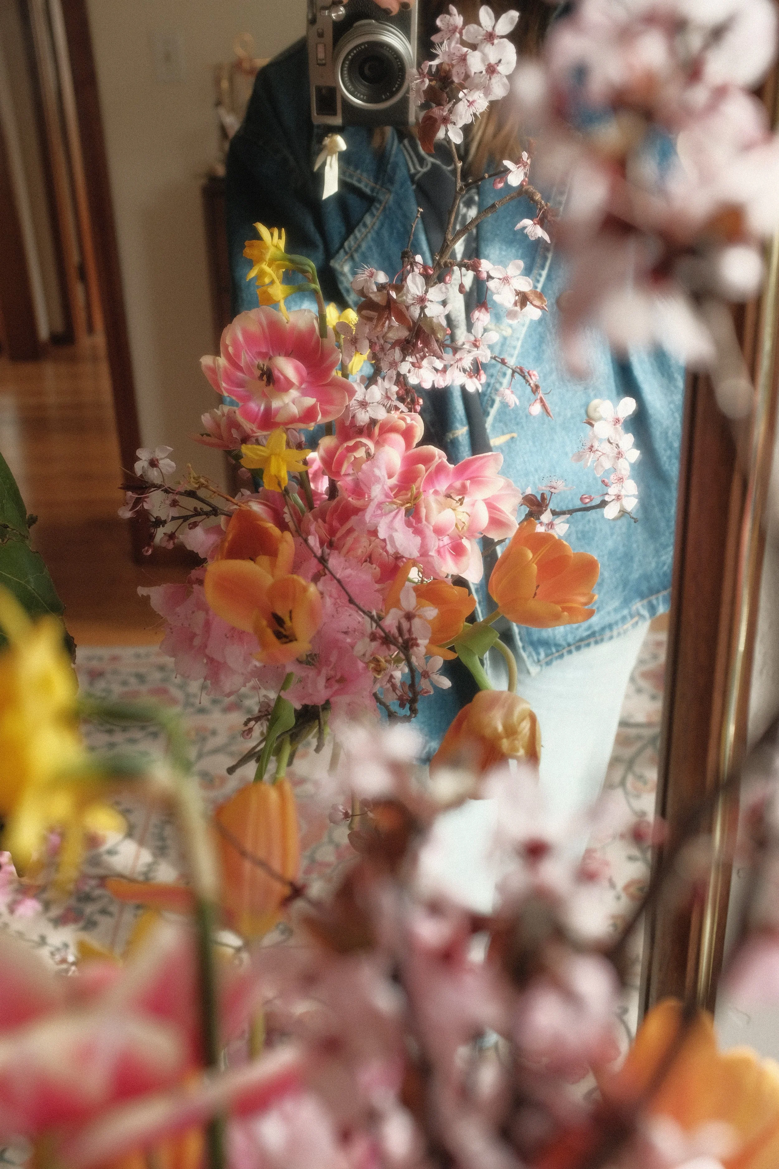 A person taking a mirror selfie with a camera, holding a bouquet of mixed pink, yellow, and orange flowers, while wearing a denim jacket and a dark shirt in a room with wood flooring and furniture.