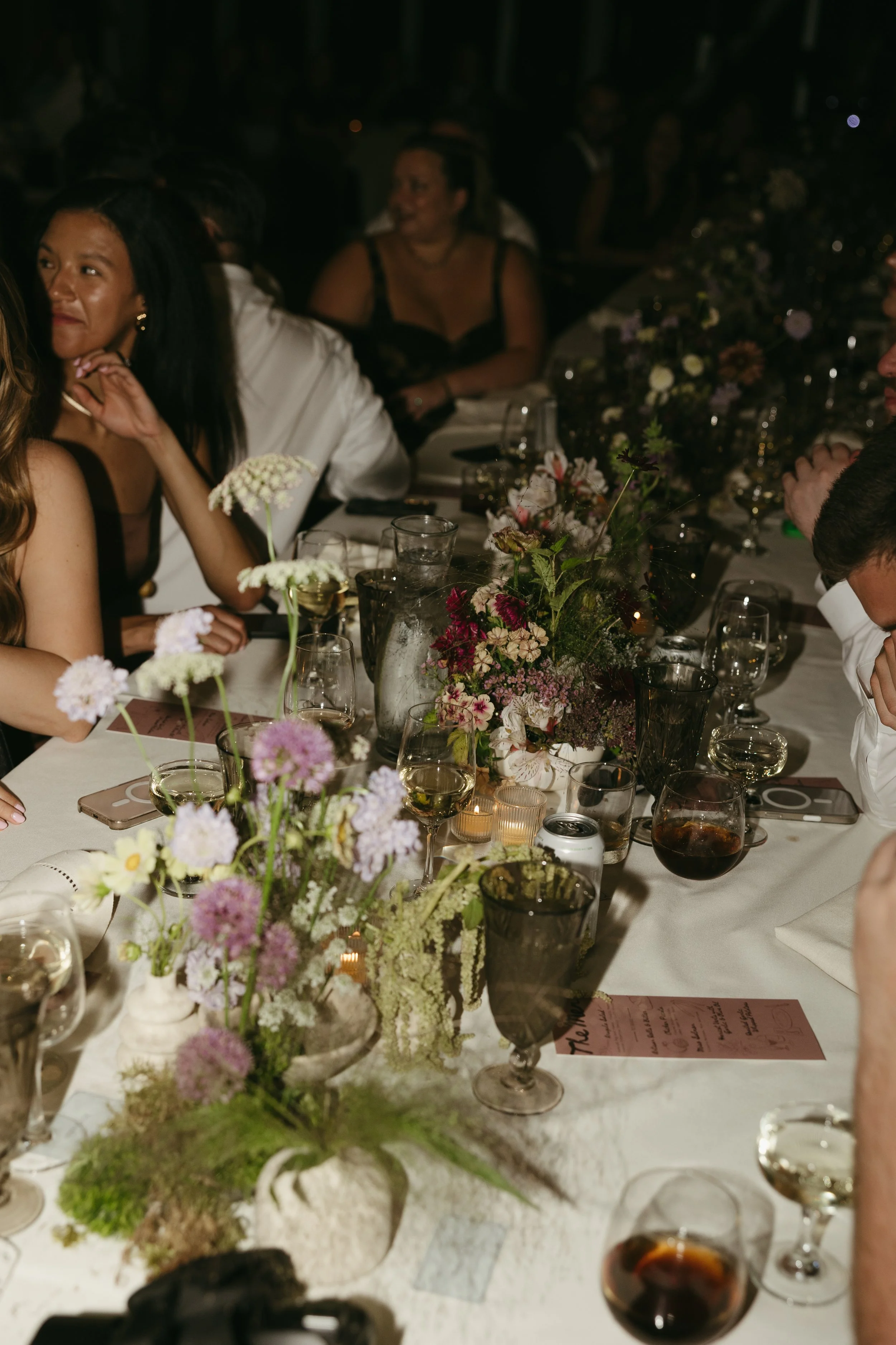 A group of people sitting at a long dinner table with floral centerpieces, glasses, and lit candles, attending a social gathering or event.