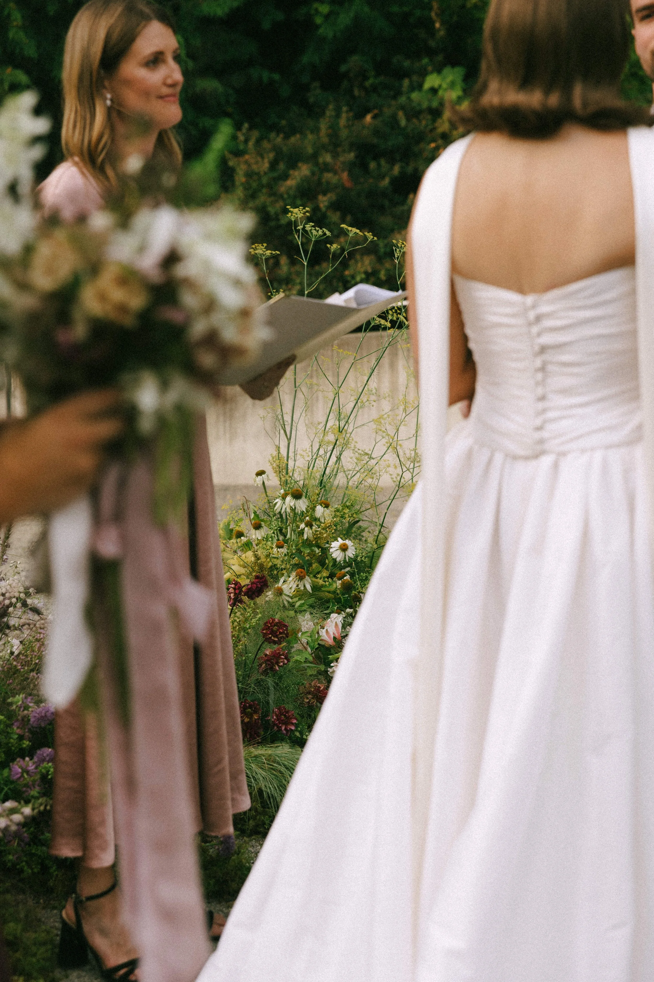A bride in a white wedding dress facing an officiant during a wedding ceremony outdoors with flowers and greenery in the background.