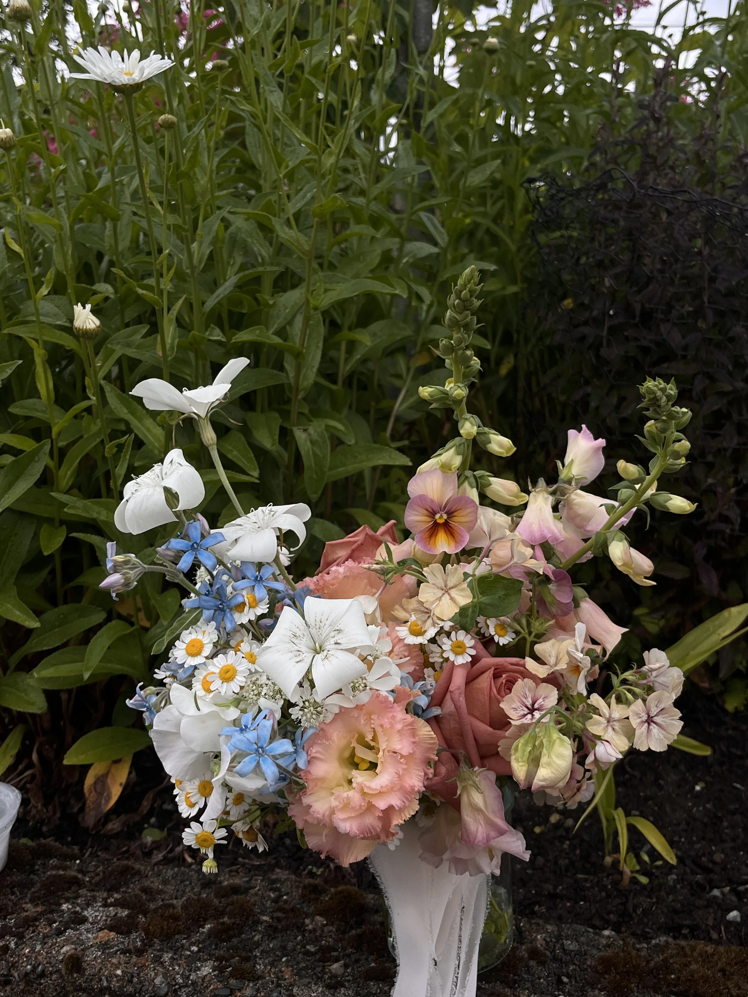 A floral bouquet with white, pink, blue, and yellow flowers in a white vase, placed outdoors on soil with lush green plants in the background.