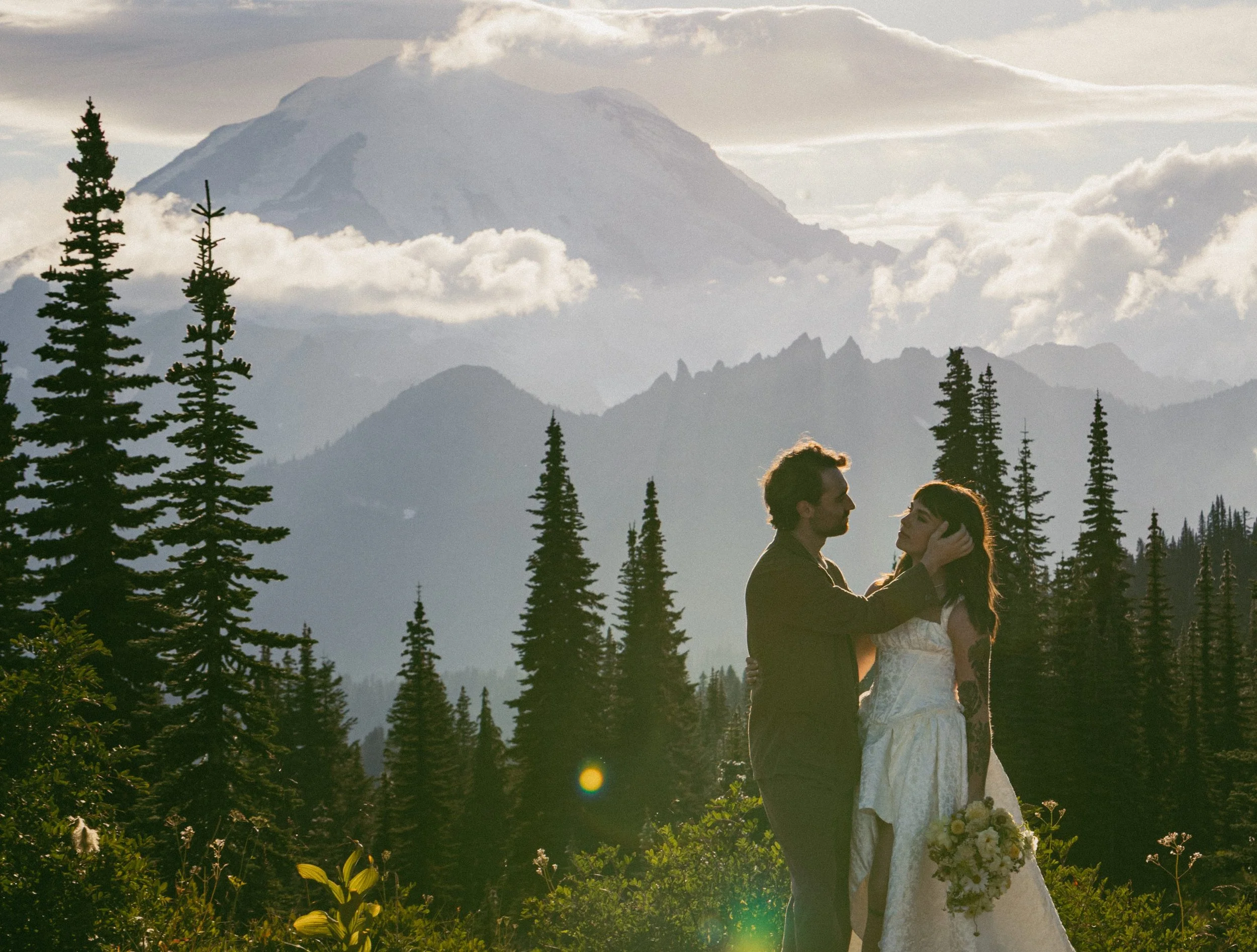 A couple holding each other in a forested area with tall pine trees and a mountain with snow in the background, during sunset.