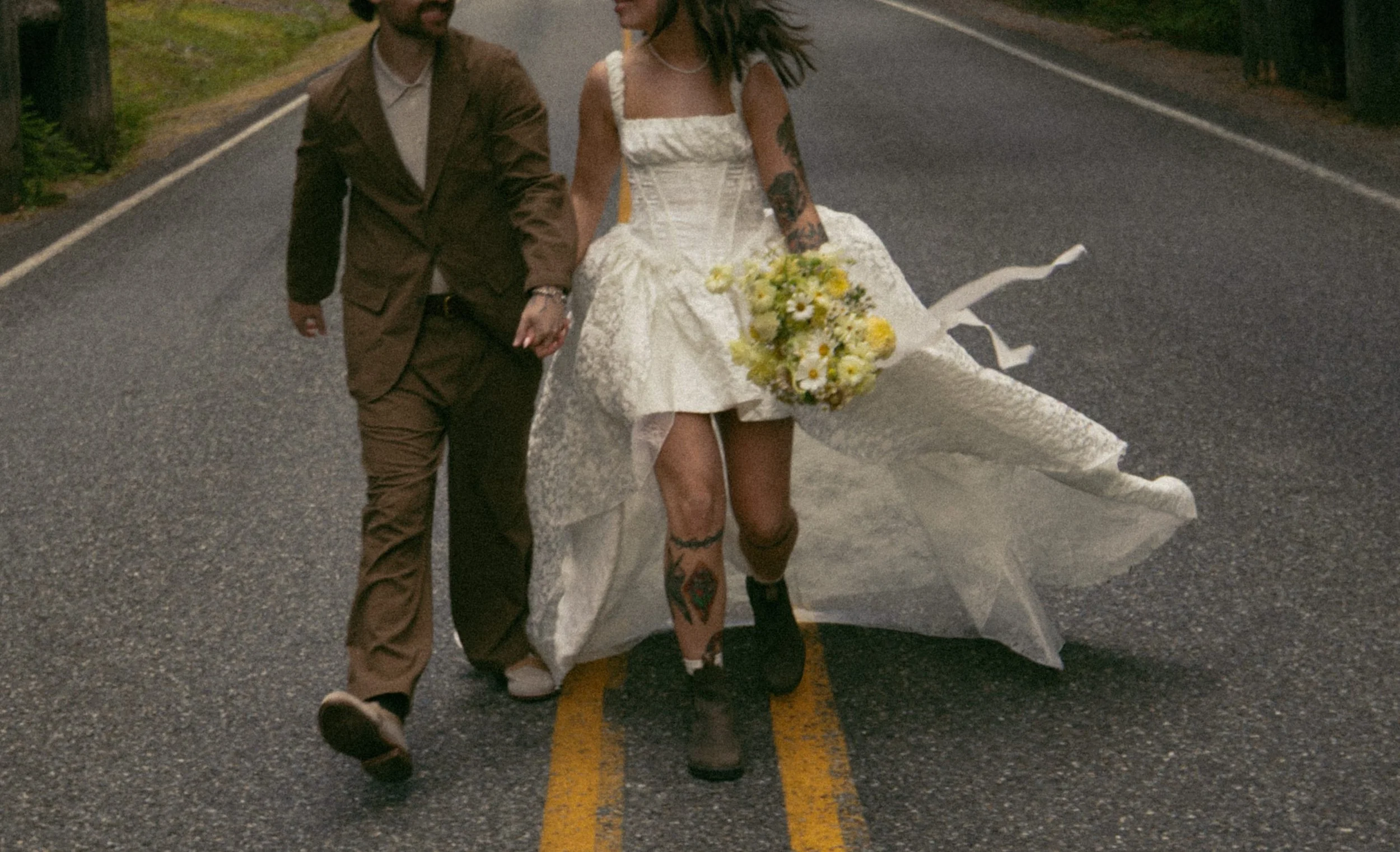 A person in a wedding dress holding a bouquet of flowers walking hand-in-hand with a person in a brown suit on a paved road.