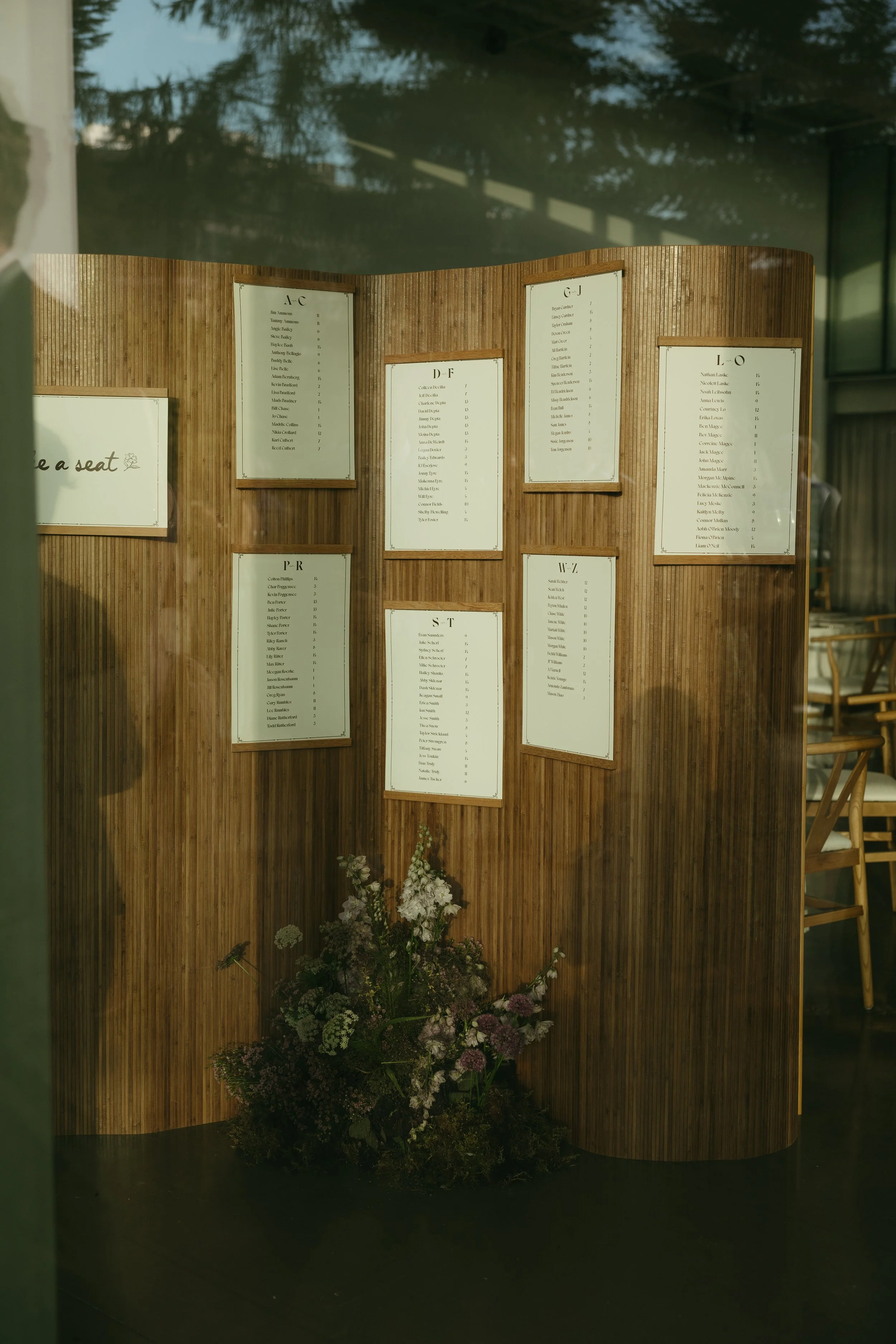 Wooden display board with various menus for a restaurant or cafe, including sections labeled AC, D-F, G-J, L-O, P-R, S-T, and W-Z, with flowers at the base.