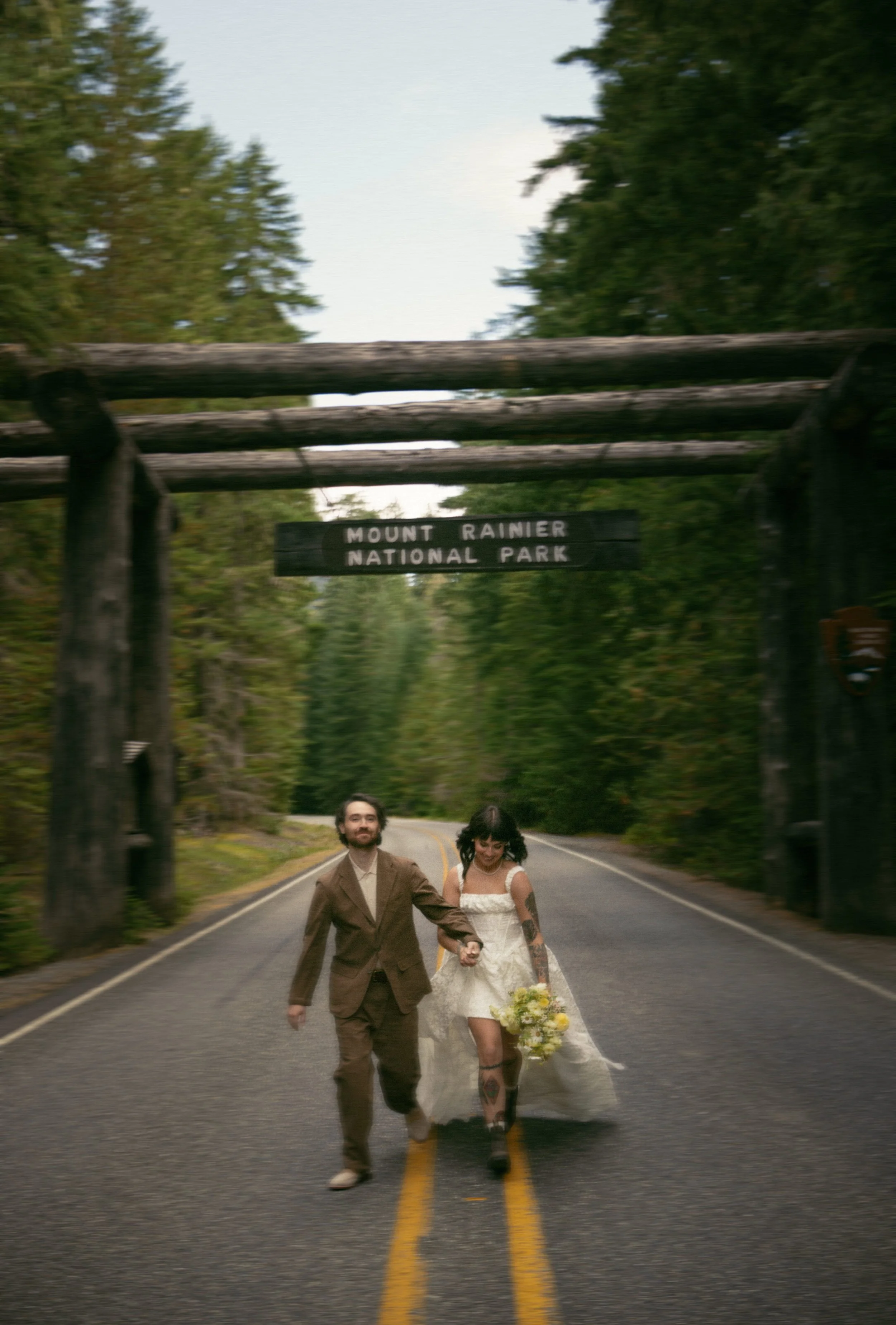 A couple in wedding attire walking on a road through Mount Rainier National Park, holding hands and smiling, with lush green trees around them.