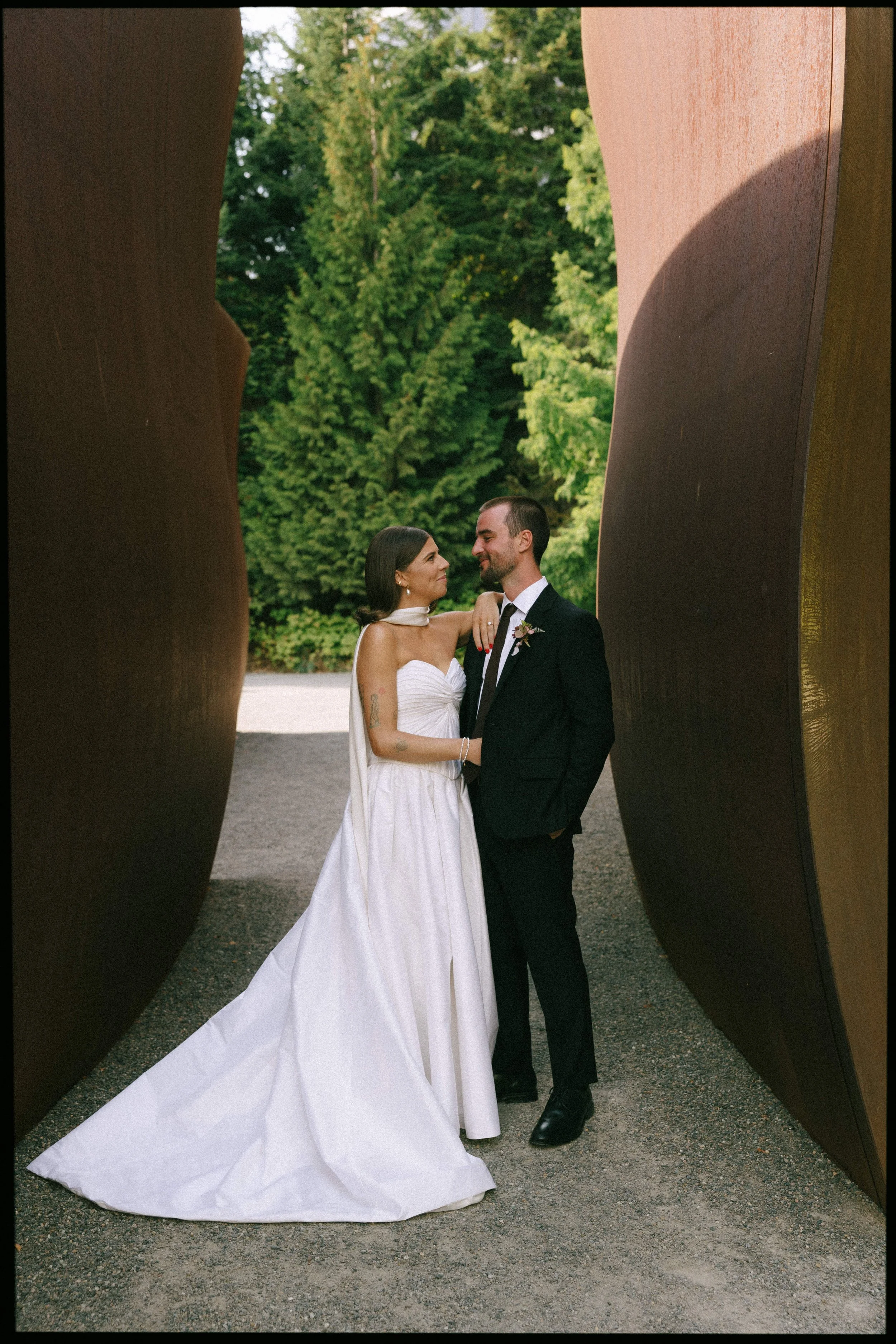A newlywed couple posing outdoors among large, curved metal sculptures with green trees in the background. The bride is wearing a white strapless wedding gown with a train, and the groom is in a black suit with a white shirt and black tie. They are l