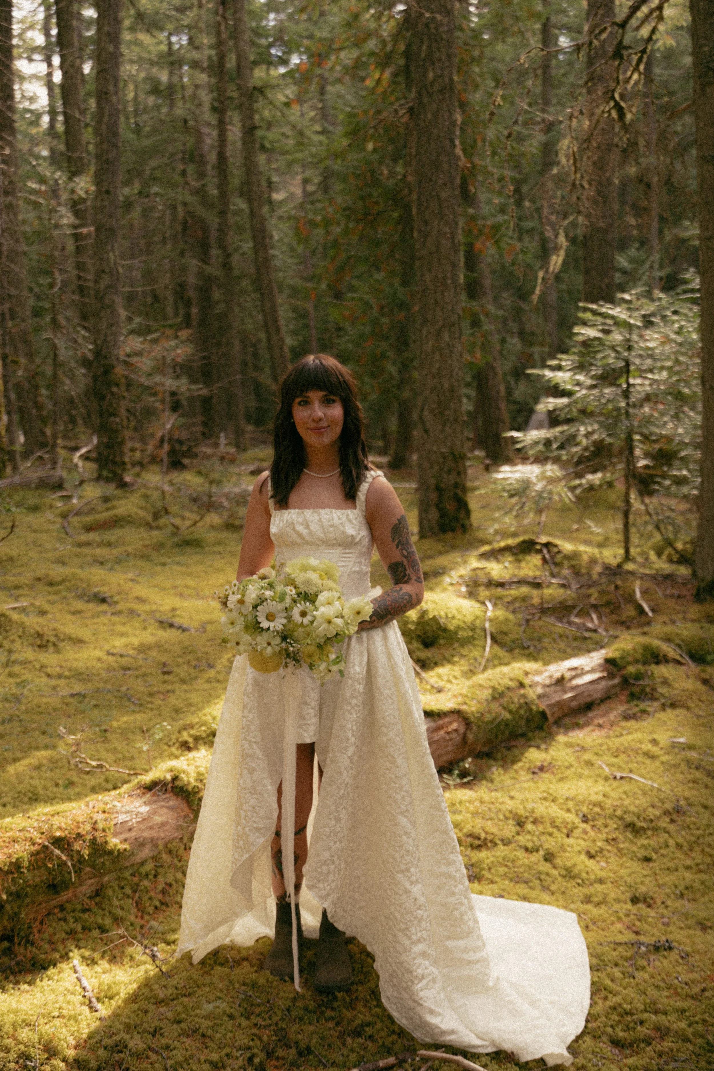 A woman in a cream-colored dress holding a bouquet of white and yellow flowers standing in a forest with moss-covered ground and tall trees.