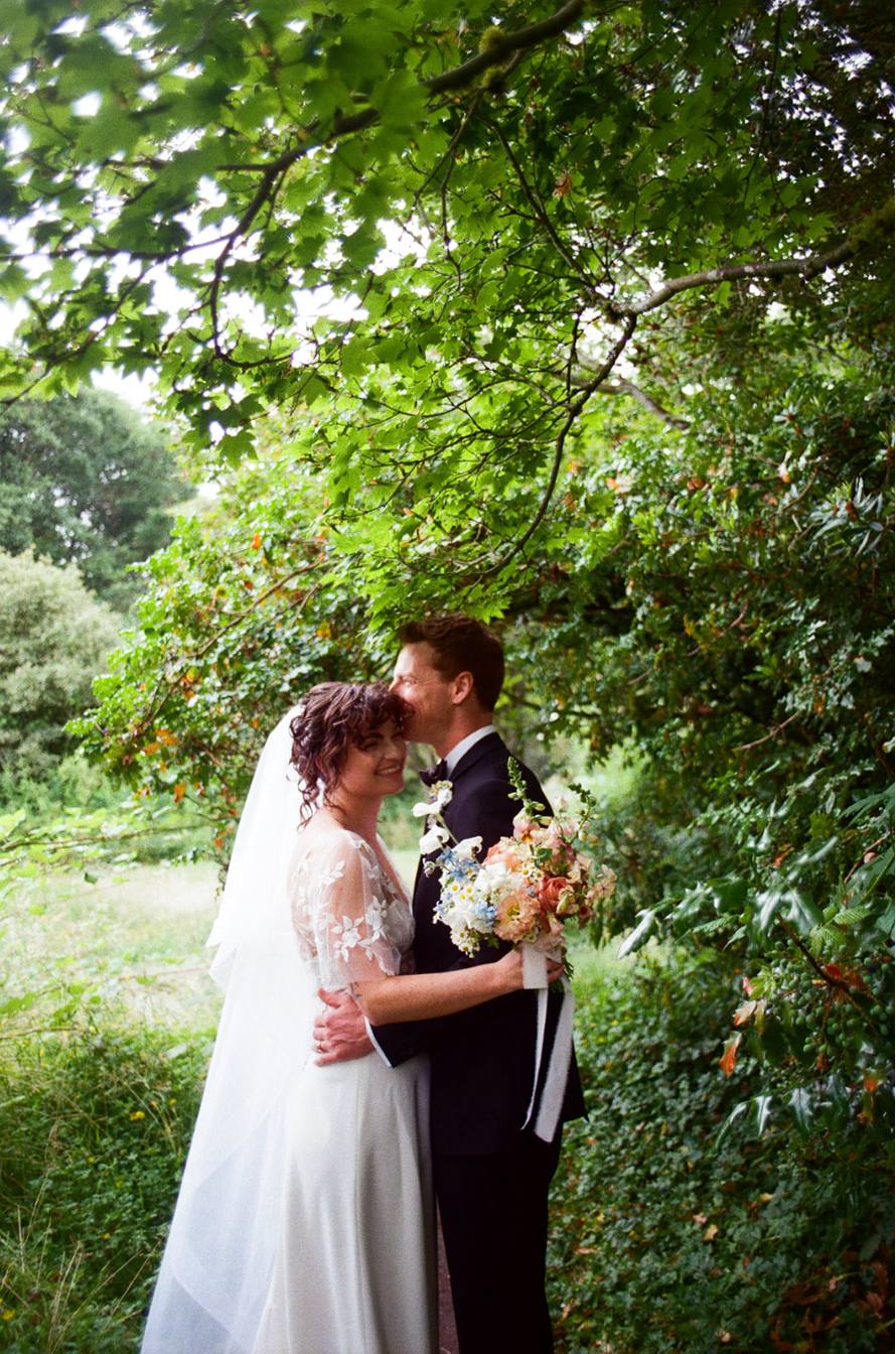A happy bride and groom embracing under green tree branches in a garden.