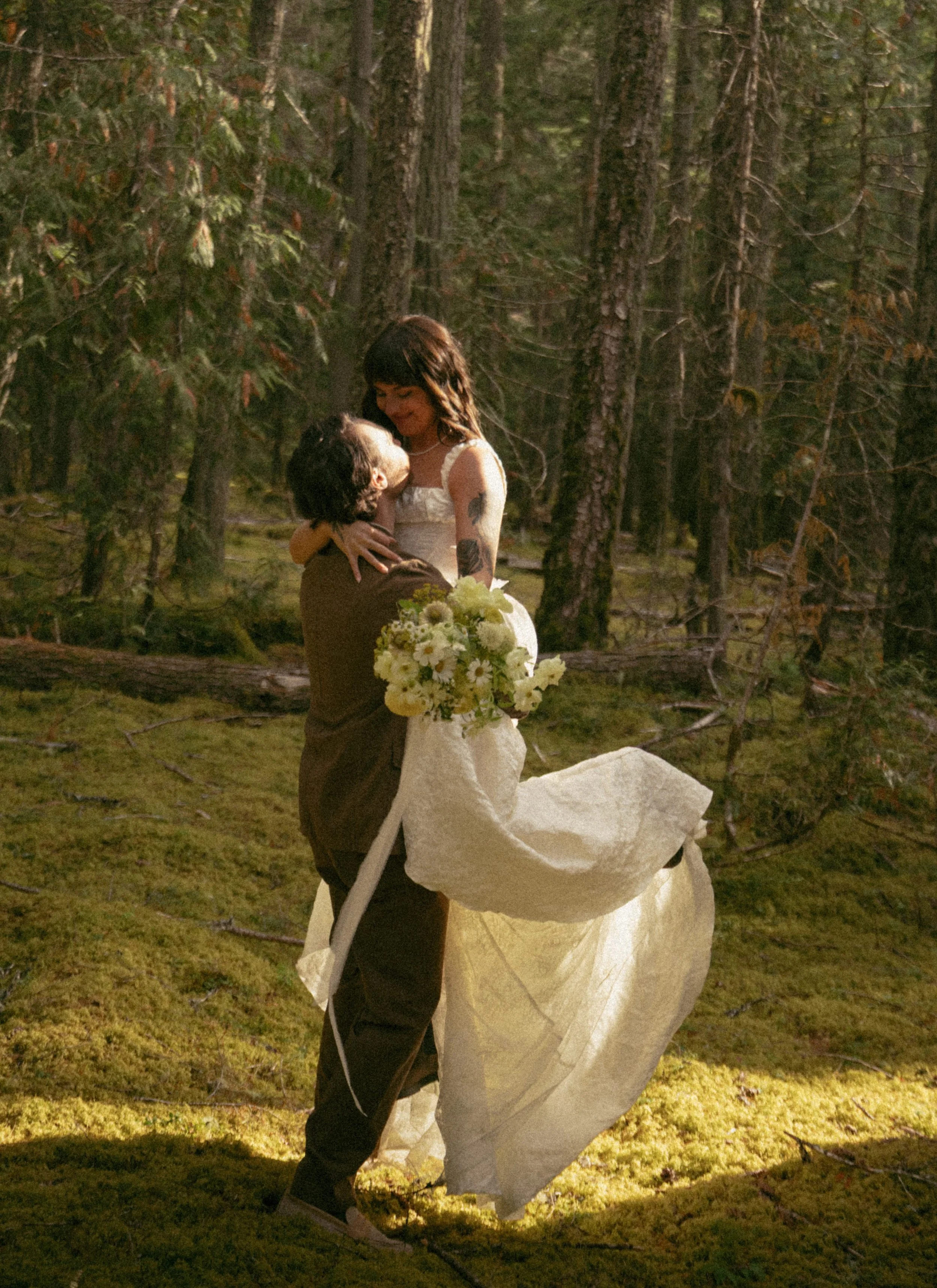 A couple embracing in a forest, the woman wearing a white wedding dress and holding a bouquet of flowers.