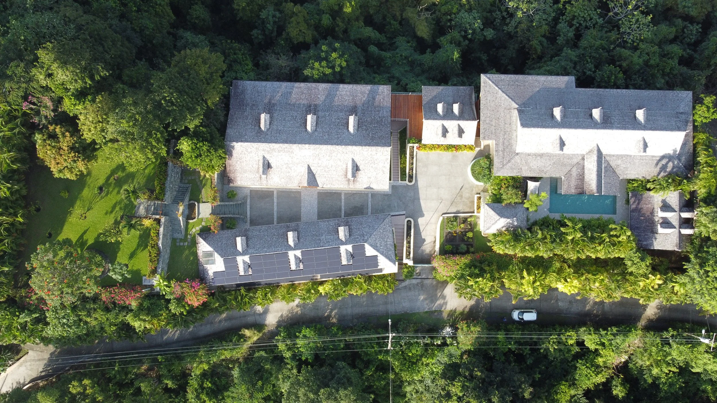 Aerial view of a large house with a swimming pool, surrounded by lush green trees and a driveway, in a tropical neighborhood.
