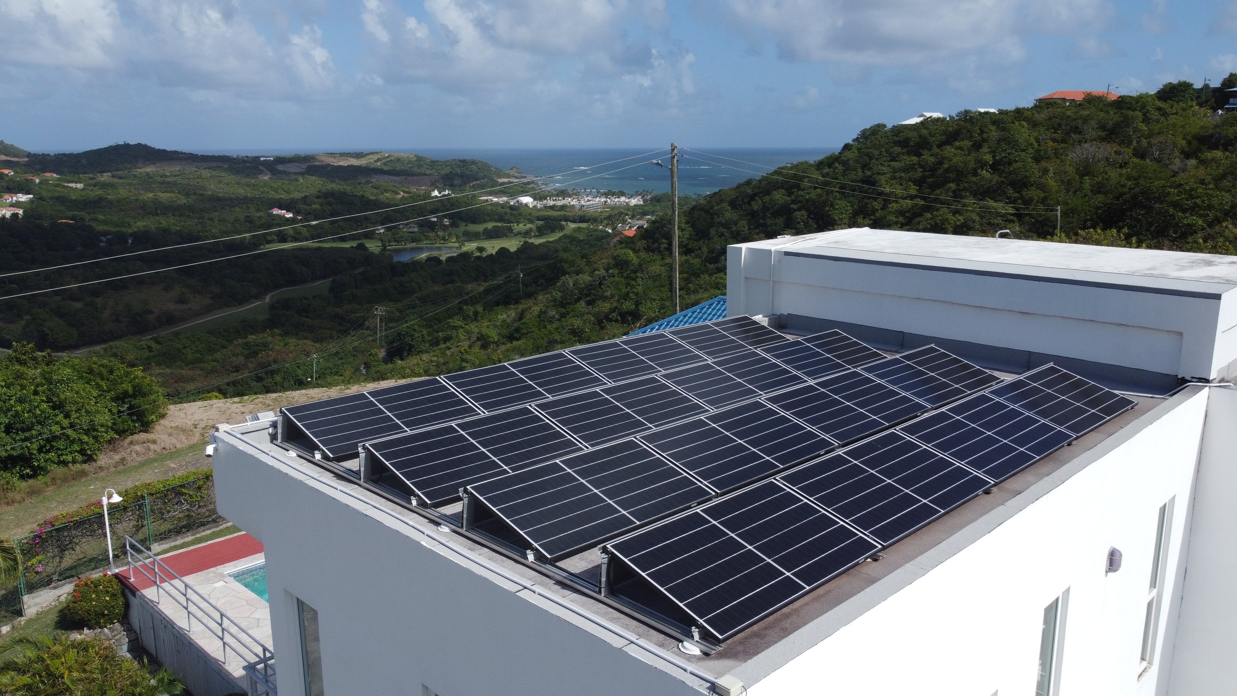 A rooftop with solar panels in a residential area overlooking a lush, green valley and the ocean in the distance.