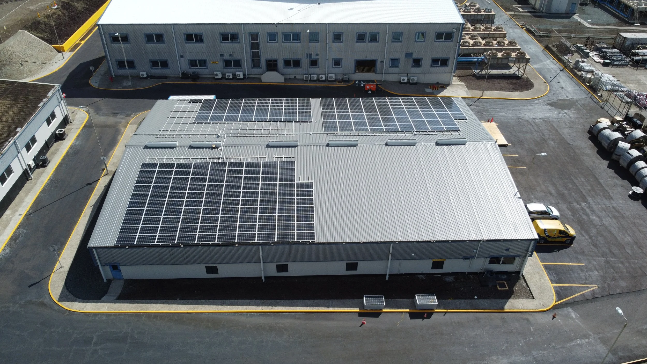 An industrial building with solar panels on the roof, surrounded by parking lot and other structures.