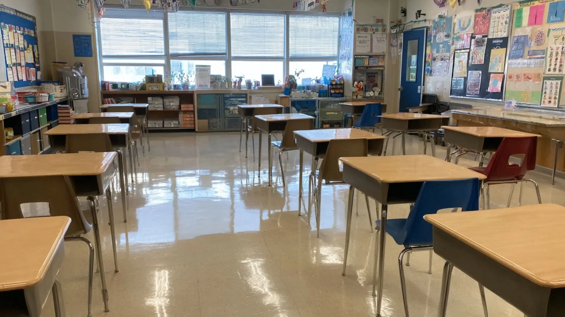 Empty classroom with wooden desks and colorful chairs, classroom decorations on the walls, large windows letting in natural light.