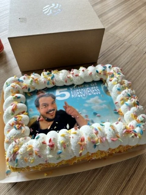 Milk is a celebratory 5 year anniversary cake with colorful sprinkles and white frosting, with a background of blue skies and clouds, placed on a wooden surface with a box nearby.