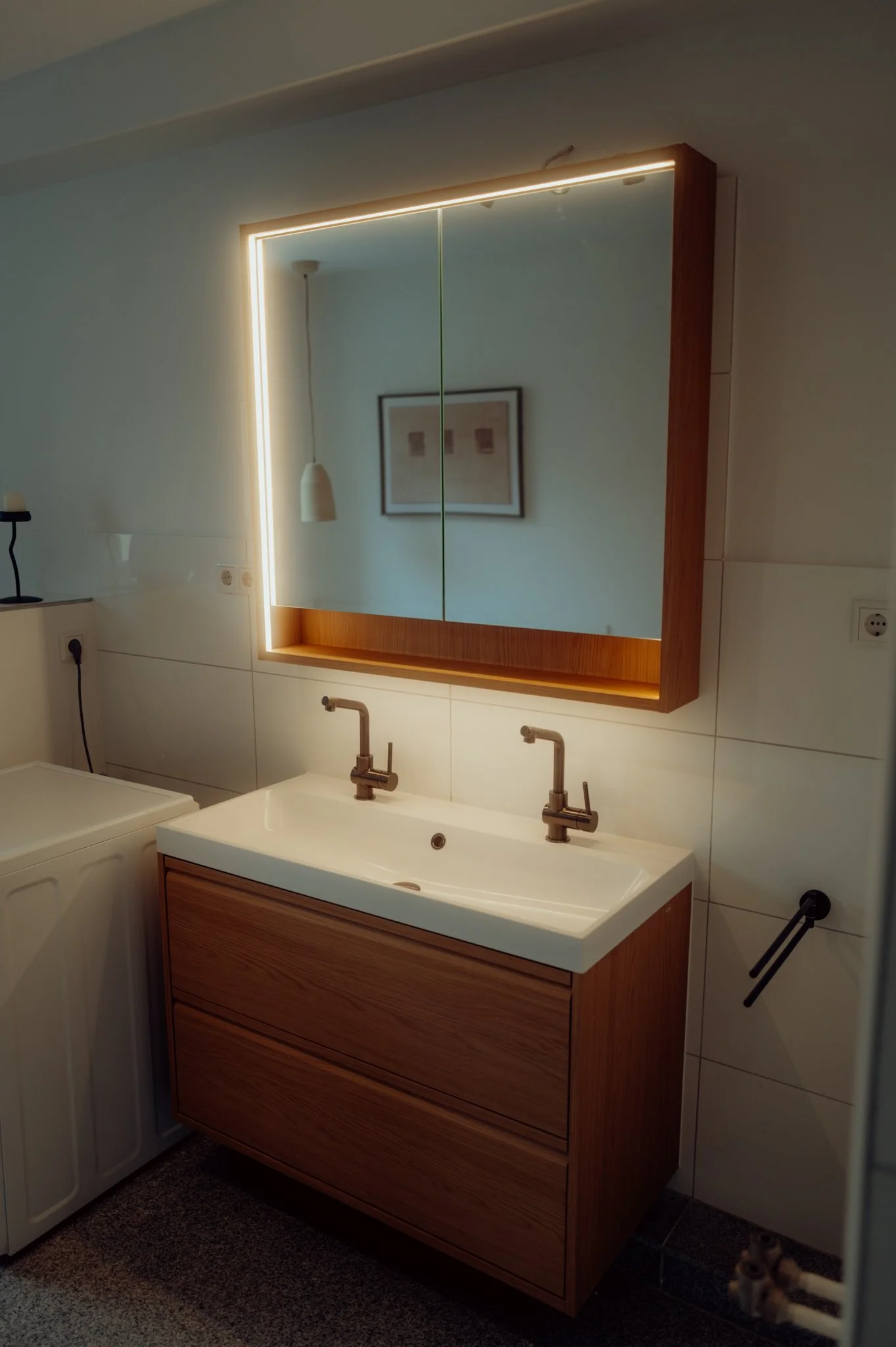 Abstract textured artwork with vertical metallic lines framed in light wood, illuminated by ceiling lights, on a white wall.