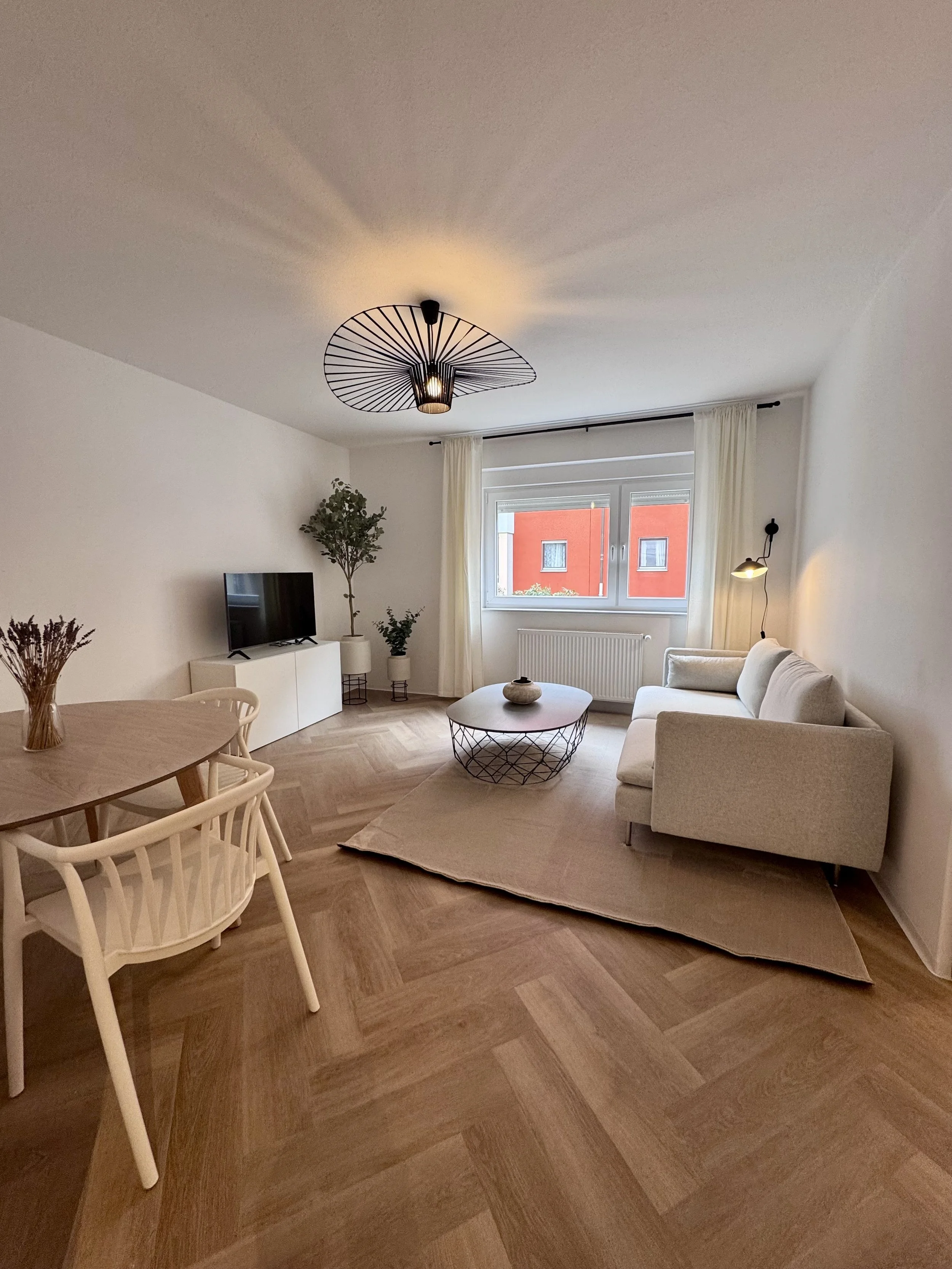Modern living room with a beige sofa, coffee table, flat-screen TV, potted plants, dining table with chairs, large window with white curtains, and wooden flooring.