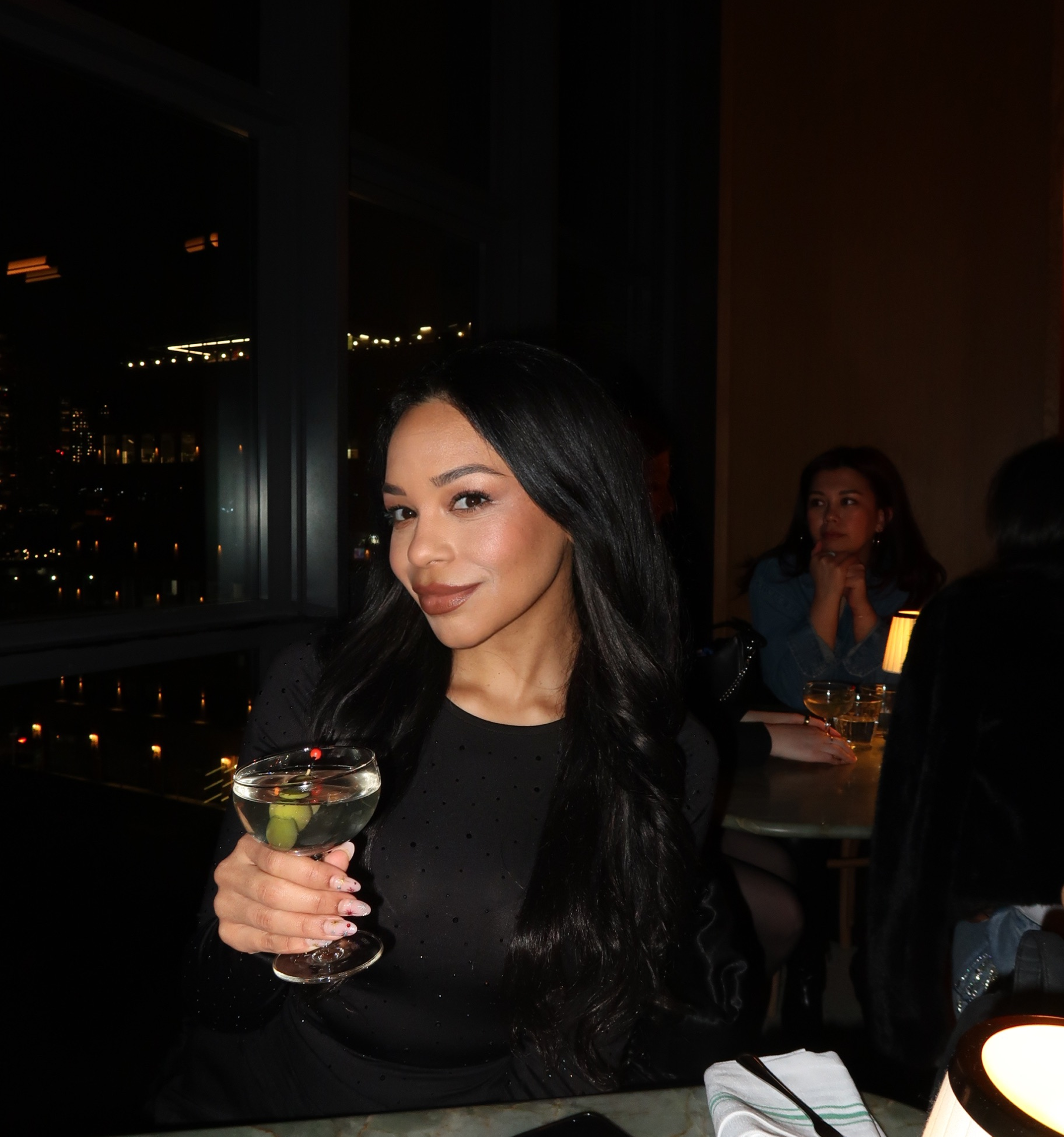 A woman with long black hair and wearing black clothing sitting at a table holding a martini glass with an olive, in a dimly lit restaurant or bar at night.
