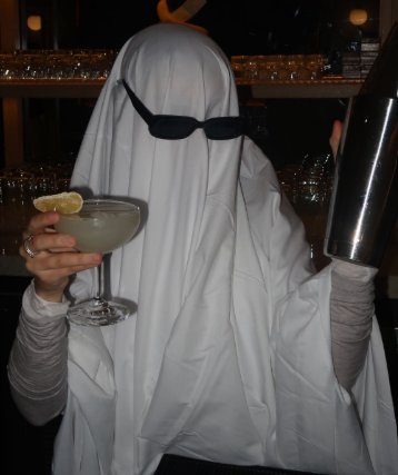 Person dressed as a ghost holding a cocktail and a cocktail shaker in a bar setting.