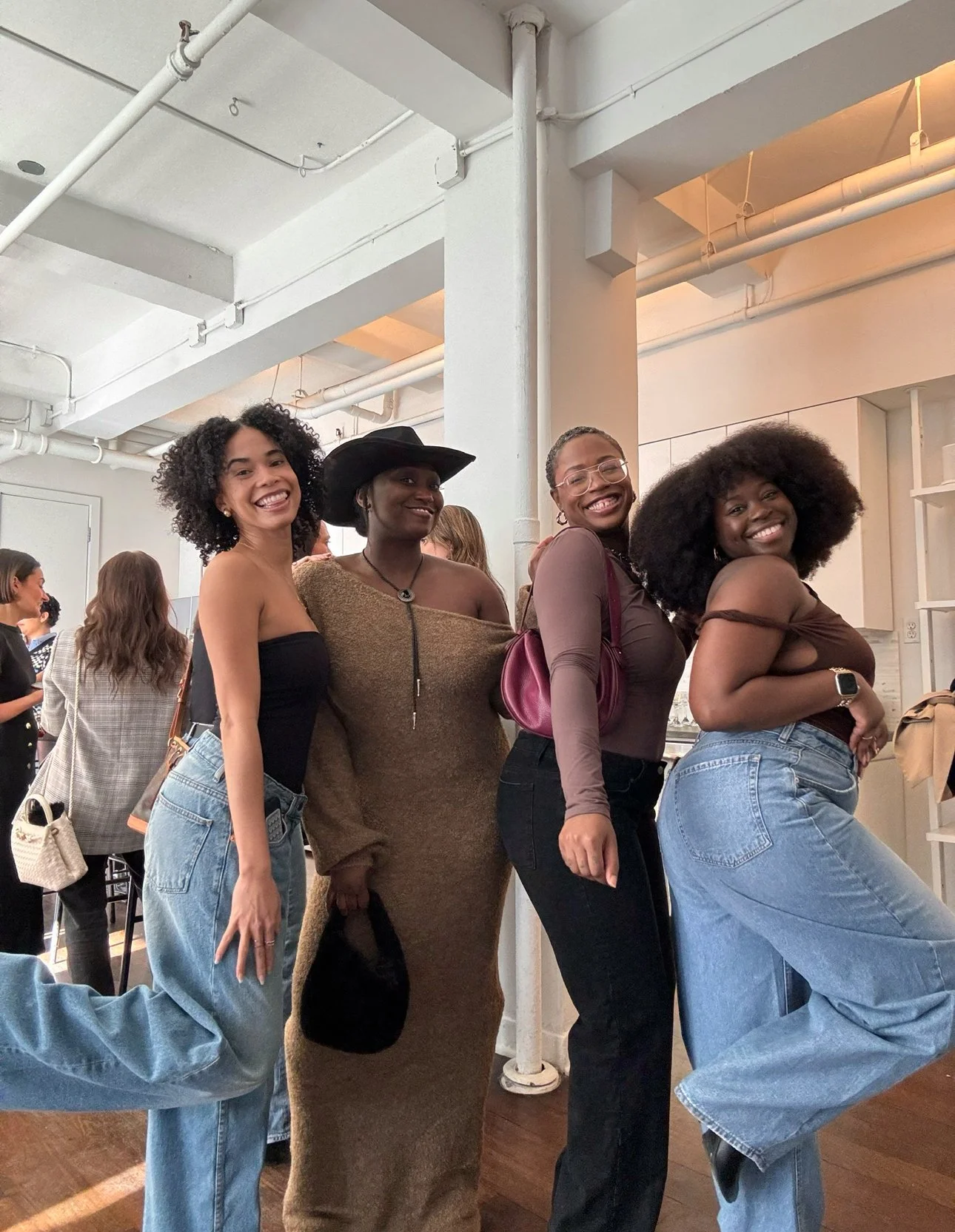 Four women standing together indoors, smiling for the camera, with a group of people in the background.