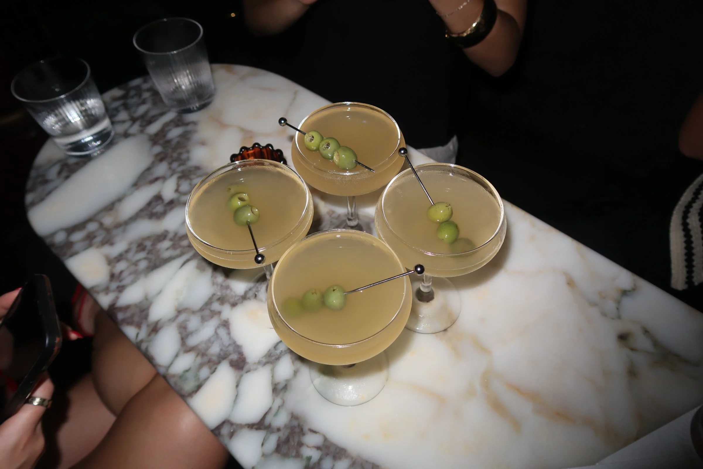 Four cocktails with green olives garnish on a marble table, with people sitting around, and glasses in the background.