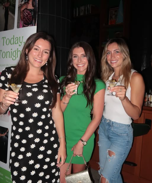Three women smiling and holding drinks at a party or gathering indoors.