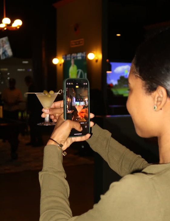 Woman taking a photo of a cocktail with a smartphone in a dimly lit bar or restaurant.