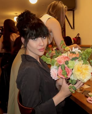 A woman with black hair and bangs holding a bouquet of pink, yellow, and orange flowers at a social gathering.