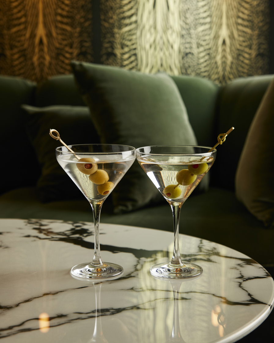 Two martini glasses with olives, placed on a marble table, with a sofa and curtains in the background.