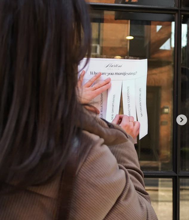 A woman peeling off a paper sign on a glass door that reads 'Start: Who are you manifesting?' with options listed underneath.