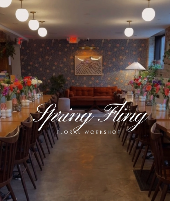 Interior of a floral workshop with tables on both sides adorned with vases of colorful tulips and other flowers, a burnt orange sofa at the back, and a blue floral wallpaper on the back wall.