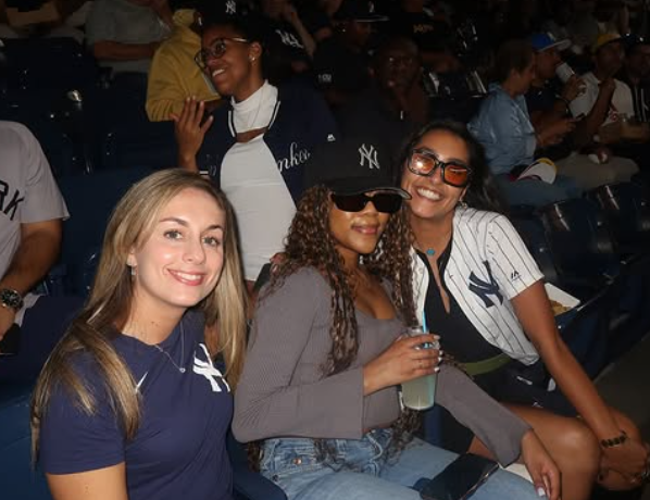 Three women sitting in stadium seats, wearing New York Yankees jerseys and apparel at a baseball game, smiling and enjoying themselves.