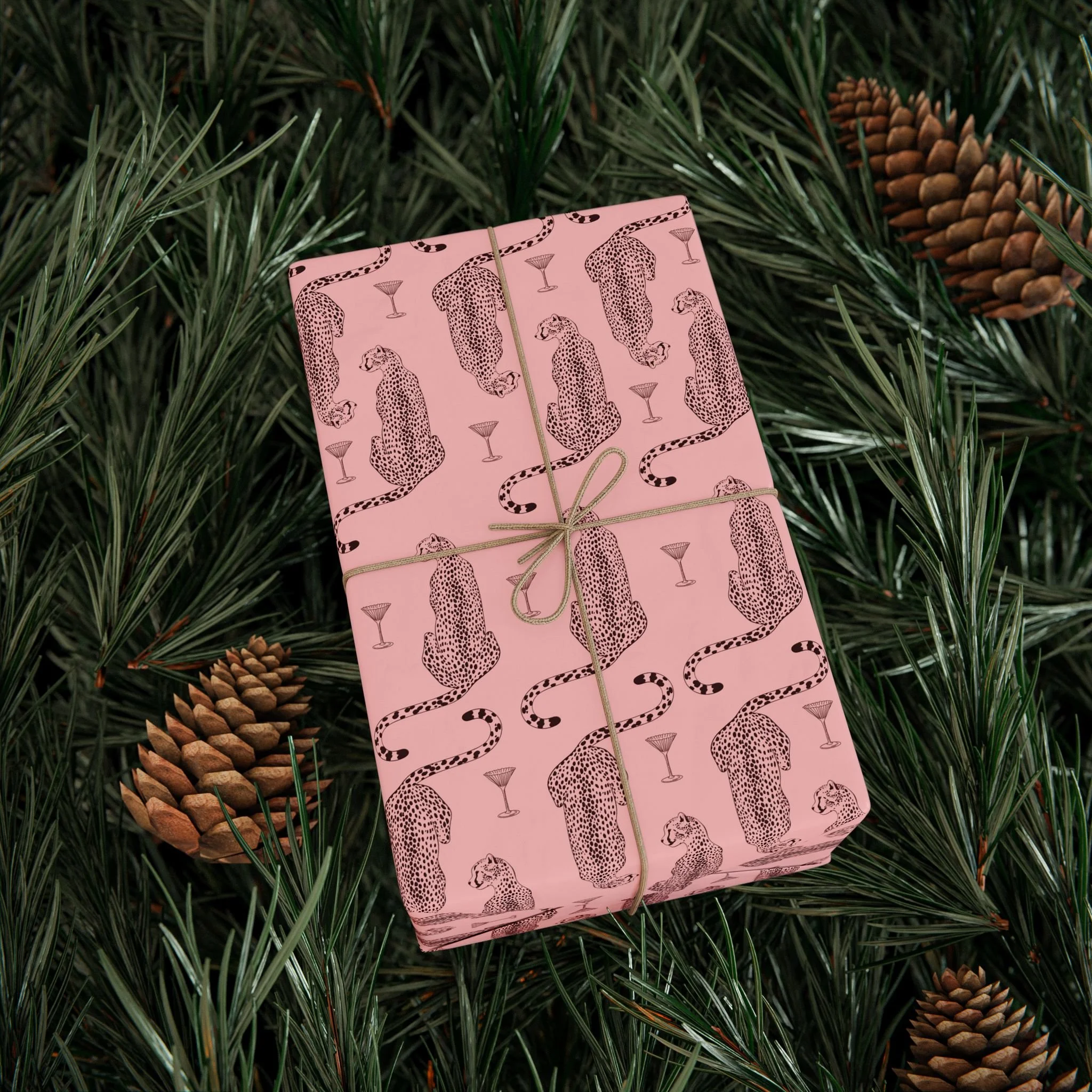 Pink gift box with black leopard print and martini glass illustrations, wrapped with a tan string, placed on green pine branches with pine cones.