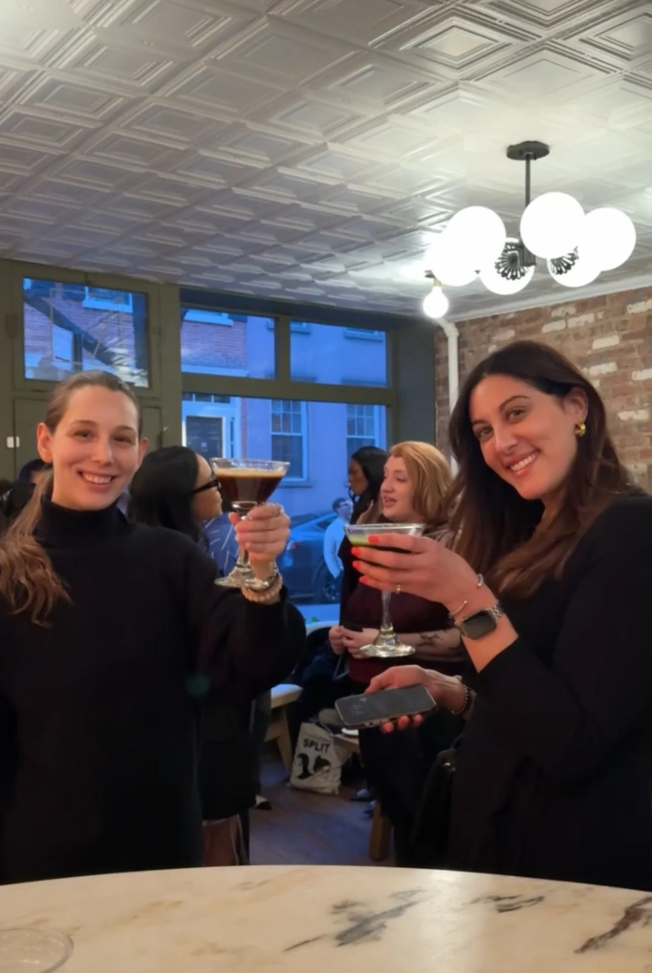 Two women smiling and celebrating with cocktails in a lively indoor setting with a brick wall, large windows, and modern lighting fixtures, surrounded by other people.