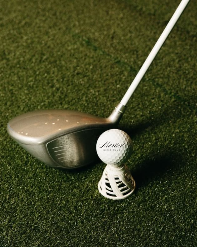 A golf club, a golf ball with 'Martini Girls Club' written on it, and a white golf tee are placed on a green putting surface.