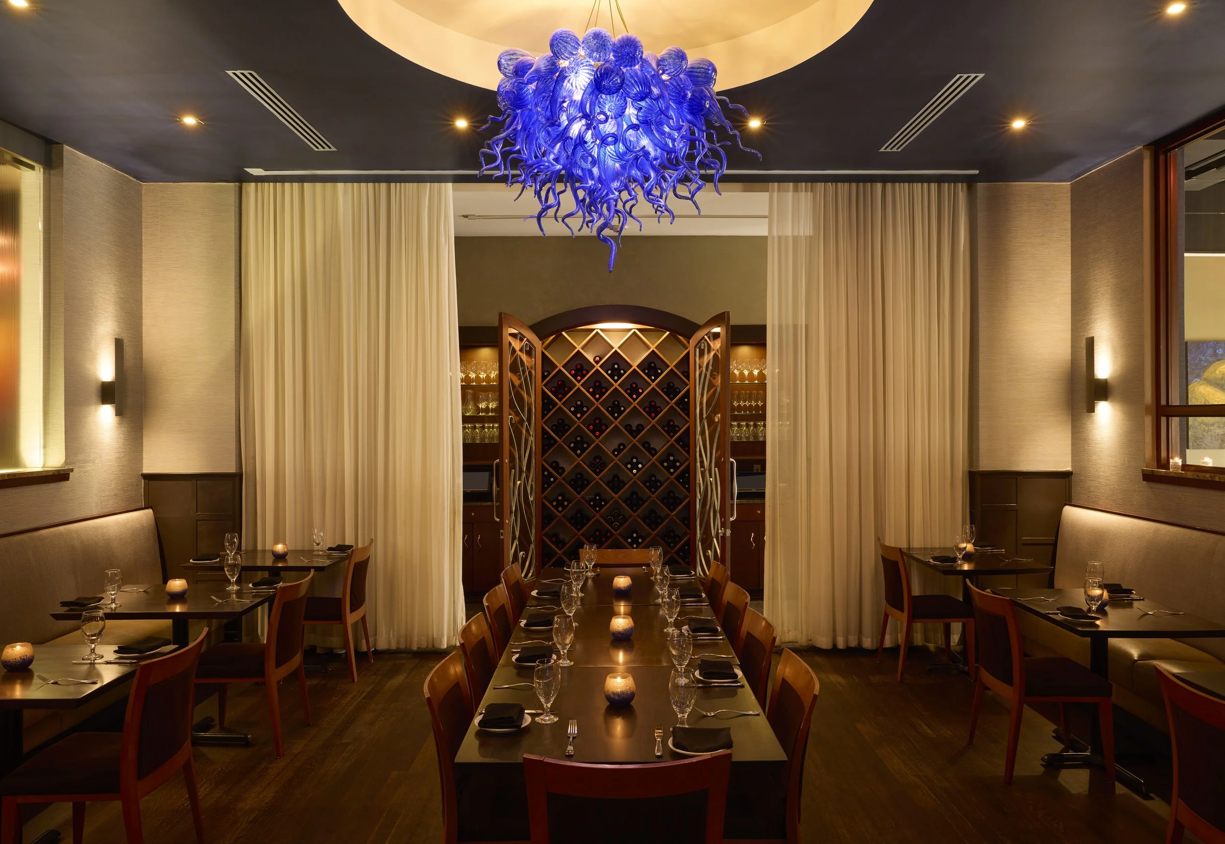 Sophisticated private dining area with a radiant blue Chihuly-style chandelier, wine storage display, and intimate seating arrangement.