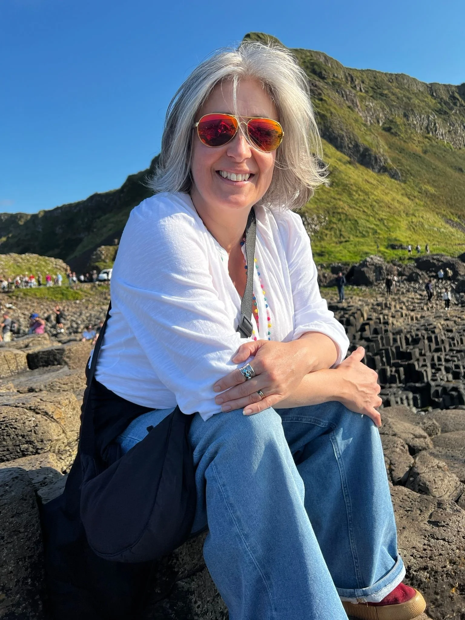 Photograph of Artist Catherine Burbridge sitting down at The Giant's Causeway. It is a sunny day and she is wearing sunglasses and smiling.