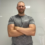 A man with a beard and short hair, smiling and crossing his arms, standing in front of a plain white wall.