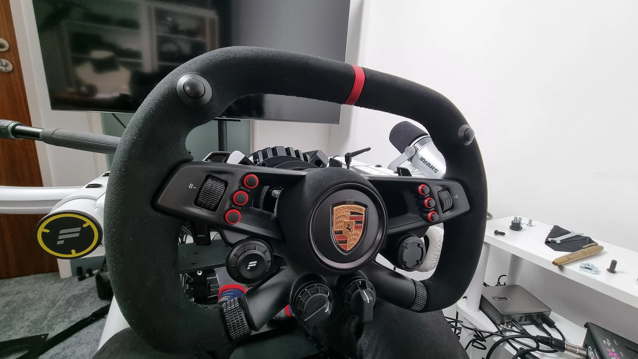 A racing simulator setup with a black Porsche steering wheel, a computer monitor, and various electronic components on a white desk.