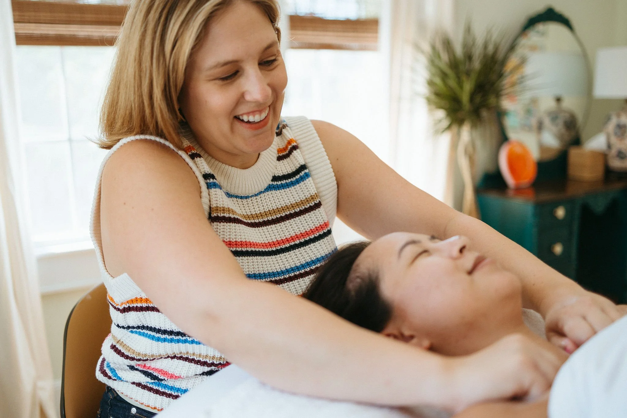 A woman with a striped sleeveless top gently holds a woman's hand during a relaxing massage in a cozy room.