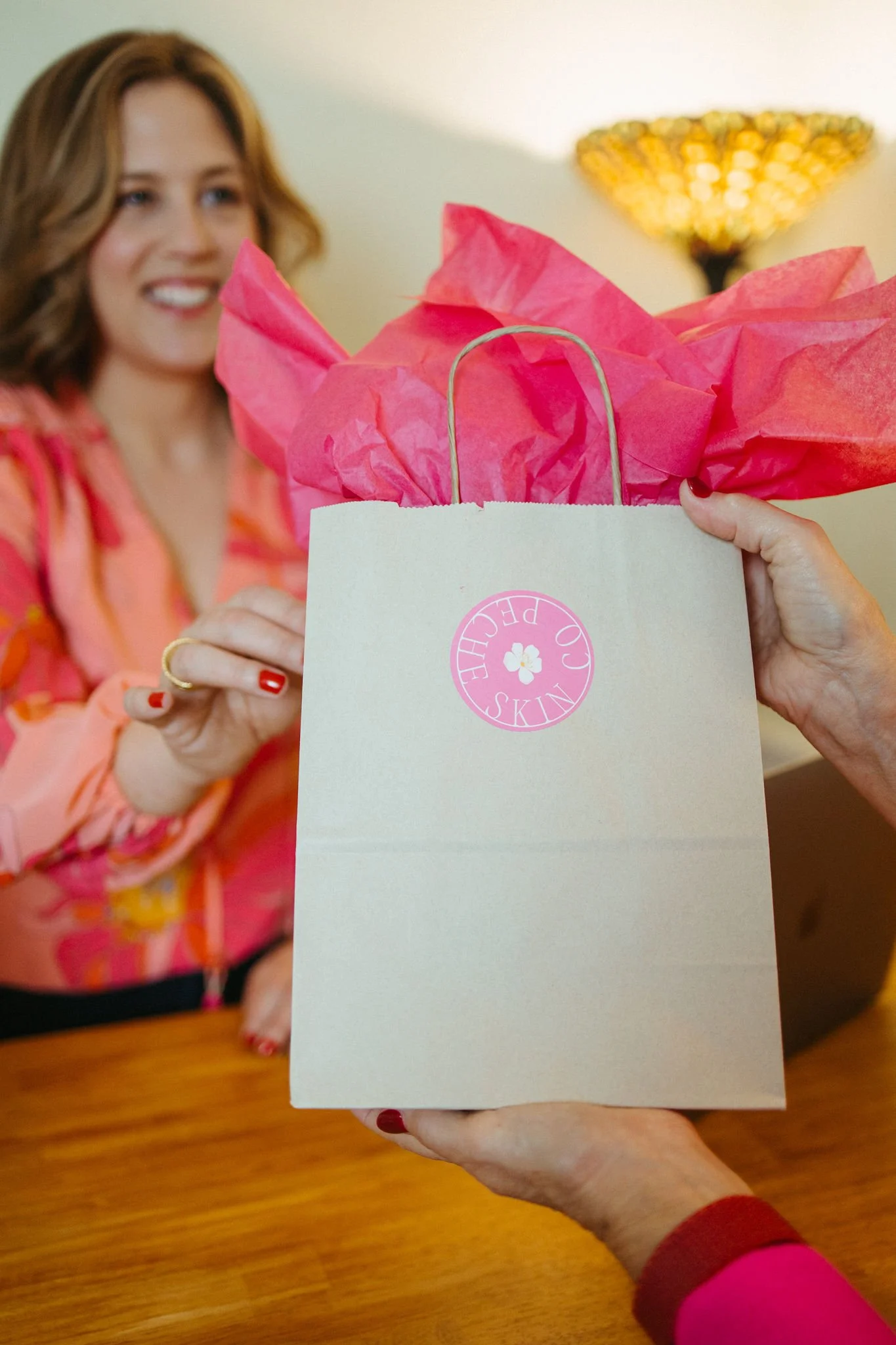 Woman with red hair smiling as she receives a gift bag with pink tissue paper from another person, in a warmly lit room.