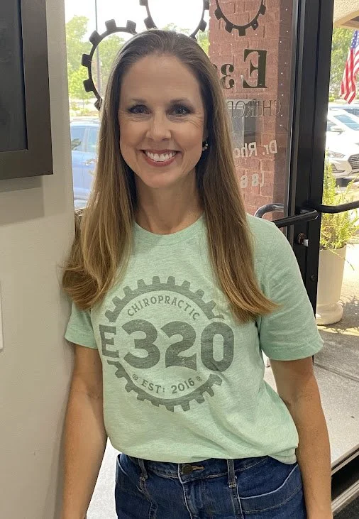 A woman with long blonde hair smiling, wearing a light green t-shirt with a chiropractic logo and the number 320, standing inside a building near a glass entrance door with an American flag and cars outside.