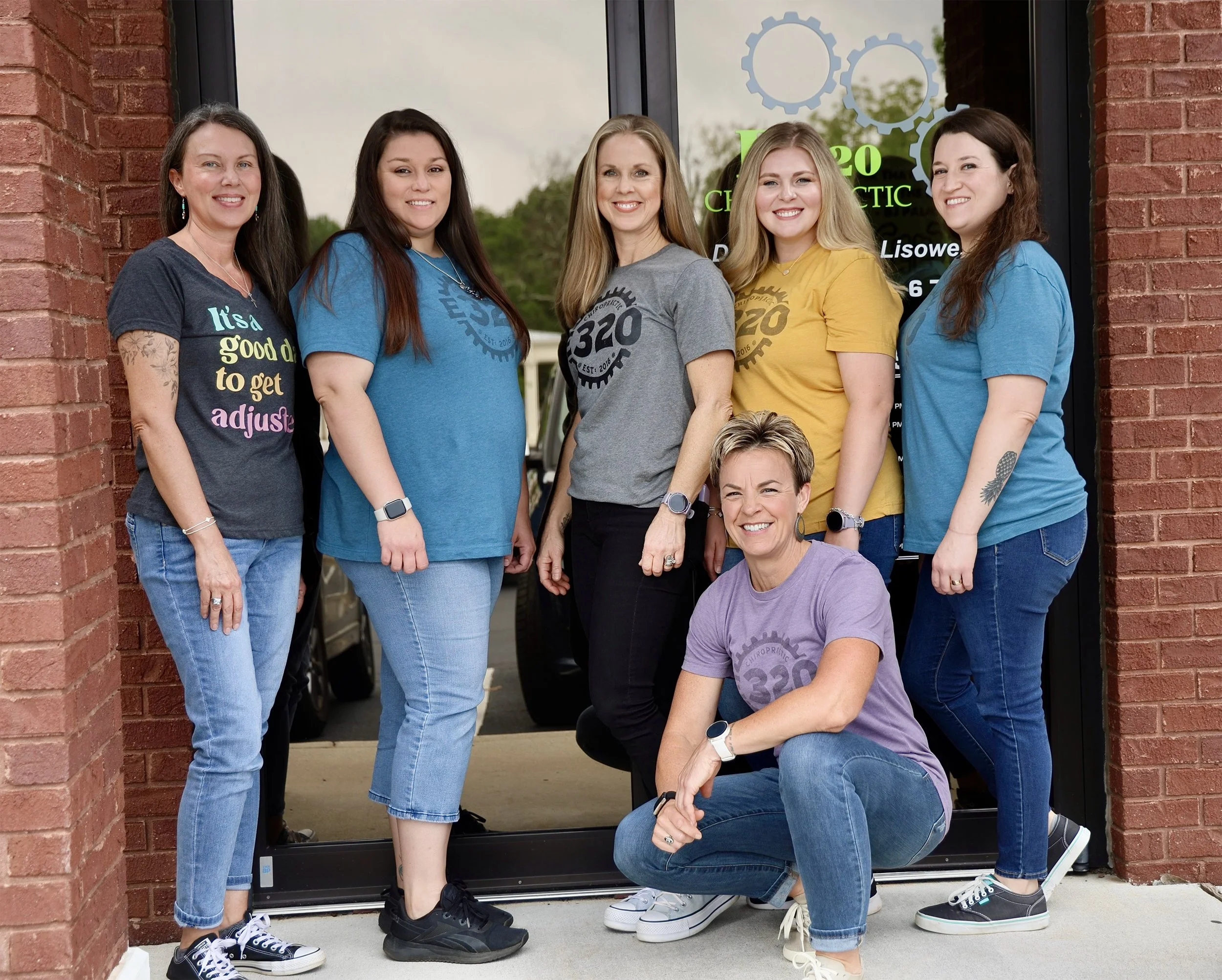 Group of five women standing together outside a building with a brick wall, smiling. They are wearing casual t-shirts with logos and jeans or pants, some with tattoos, and sneakers. One woman is kneeling in front. The building has a glass door with gear graphics and green text.