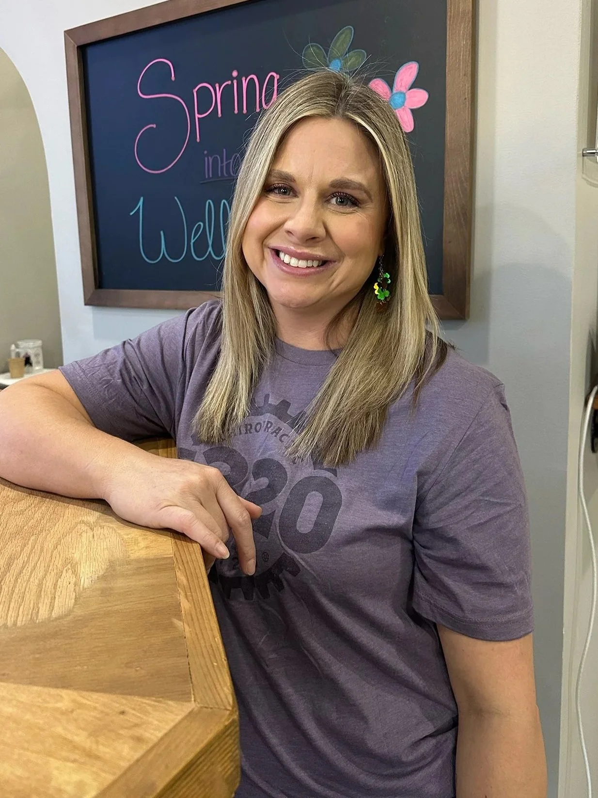 A smiling woman with blonde hair wearing a purple t-shirt with a graphic design, sitting at a wooden table with her arm resting on it, in front of a black chalkboard with colorful writing and drawings that say 'Spring into Wellness'.