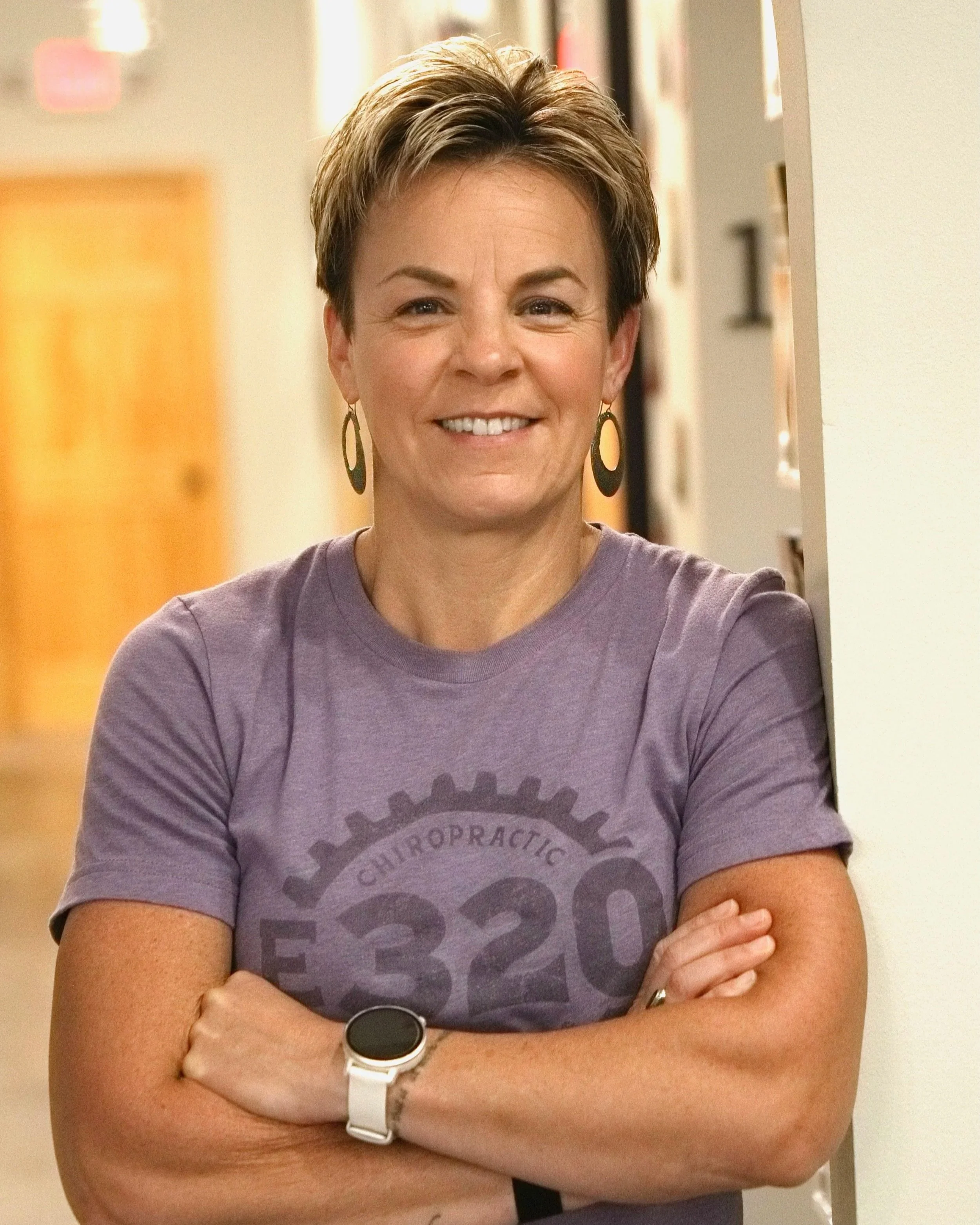 A woman with short, brown hair smiling, wearing a purple T-shirt with a gear graphic design and a transparent smart watch on her left wrist, standing with arms crossed in an indoor setting.
