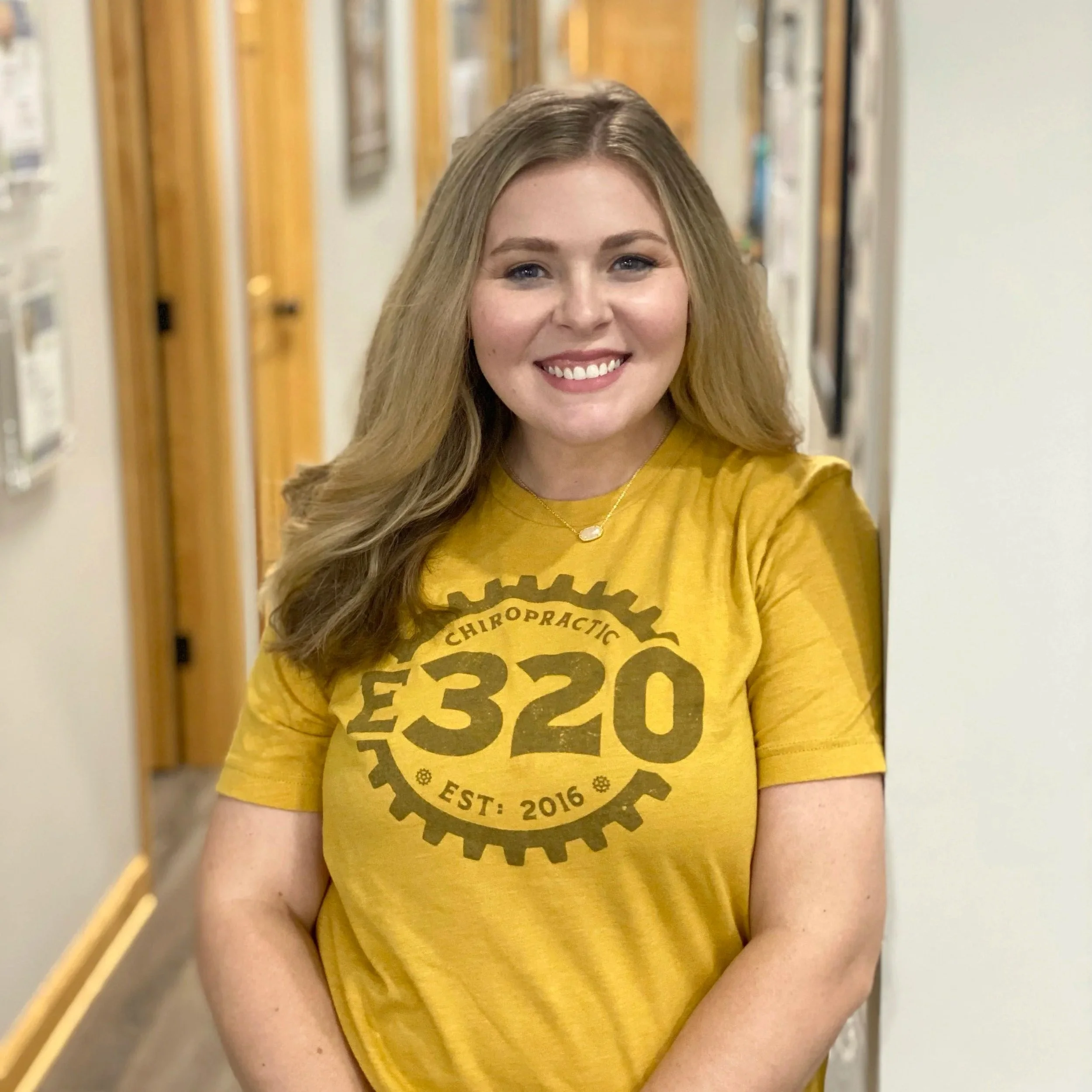 Young woman with long wavy blonde hair smiling, wearing a yellow T-shirt with black gear graphic text, standing in a hallway with wood and white walls.