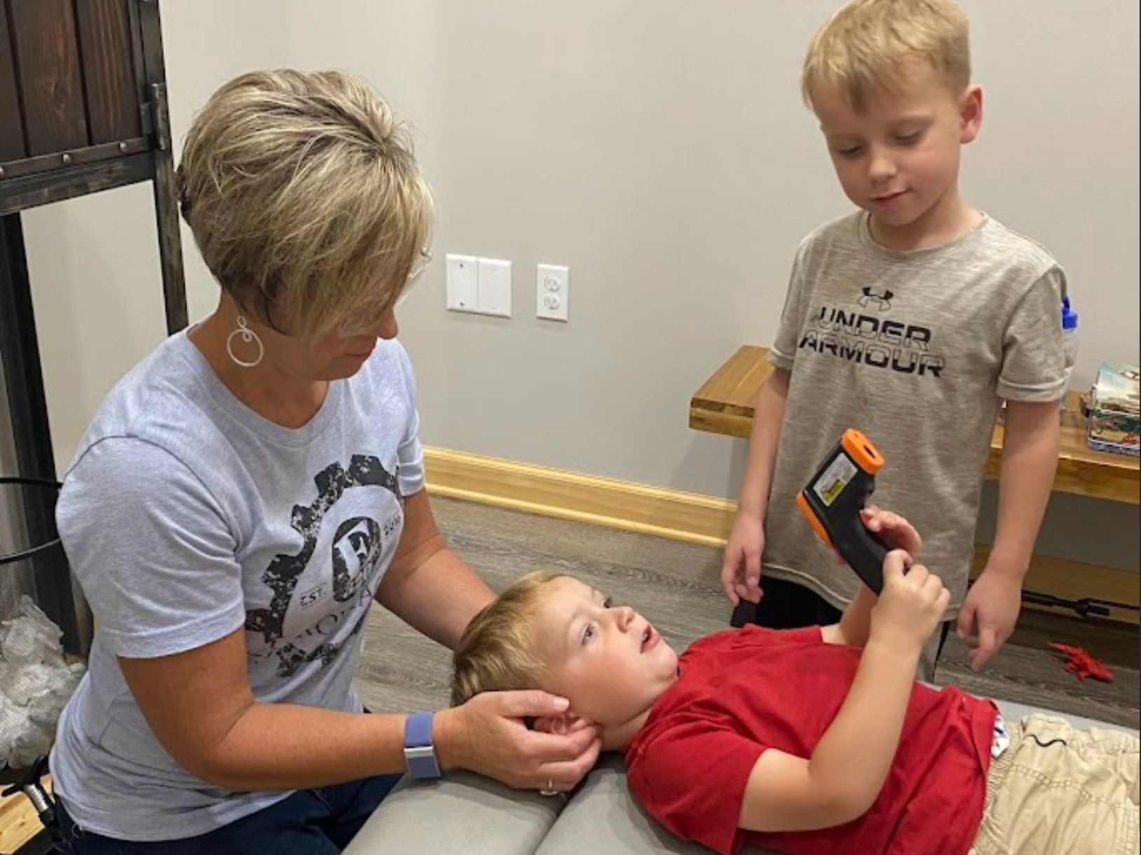A young boy lying on a table, holding a scanner device, being examined by a woman and another boy standing nearby in an indoor setting.