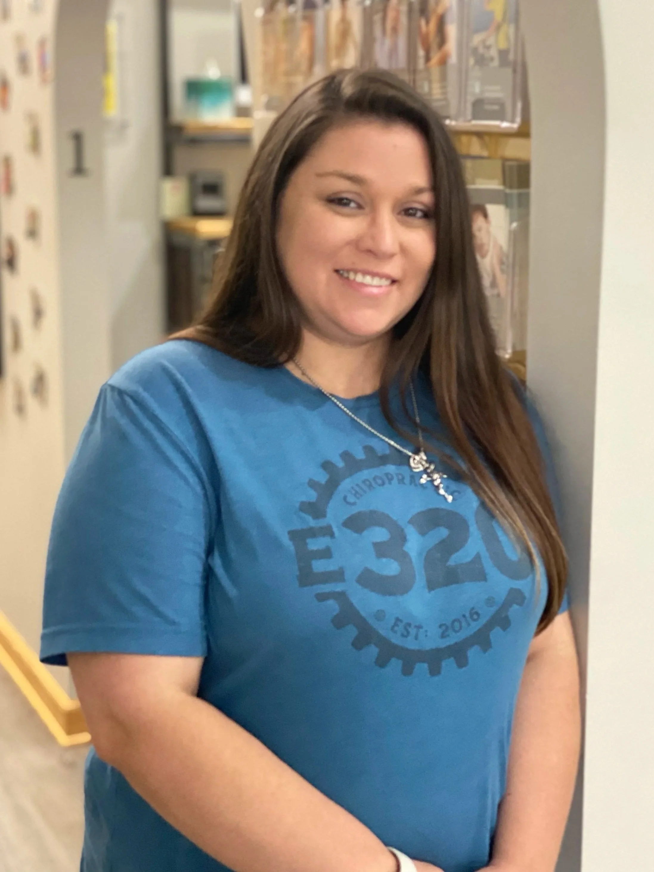 A young woman with long brown hair is standing in a hallway, smiling at the camera. She is wearing a blue T-shirt and a necklace, with a locker and some posters on the wall behind her.