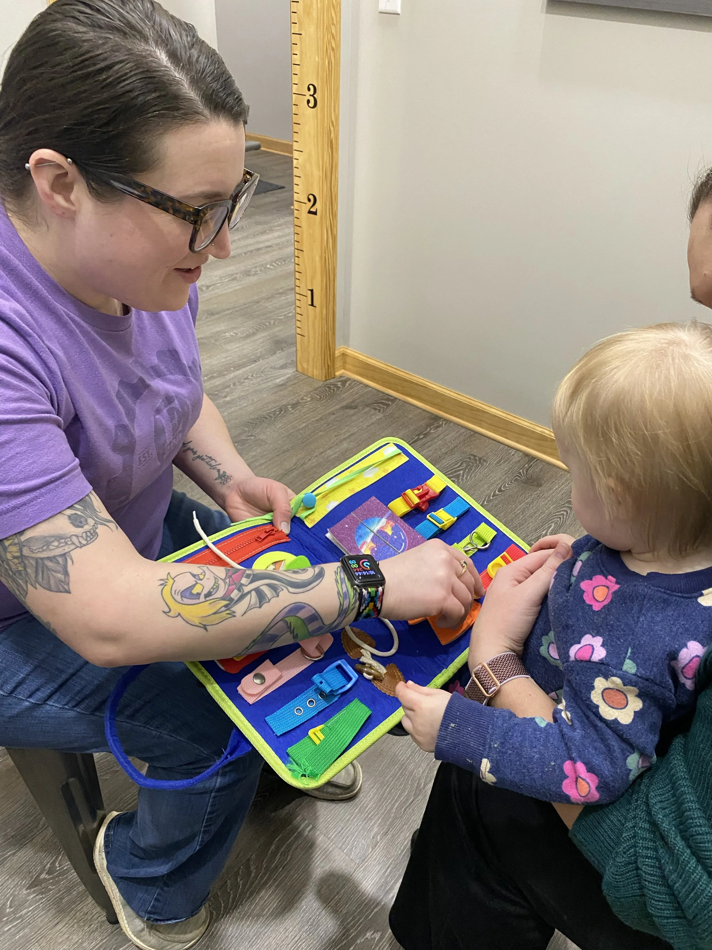 A woman with tattoos on her arms and wearing glasses is playing a game with a young girl at a table. The woman is dressed in a purple shirt, and the girl is wearing a dark blue shirt with colorful flowers. They are sitting at a blue play mat with various colorful objects on it, engaging in an activity together.